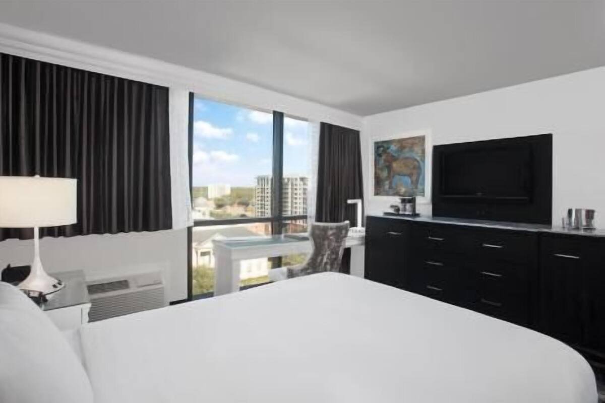 A hotel room with a large bed, a flat-screen TV, a desk with a chair, and a view of buildings through large windows.
