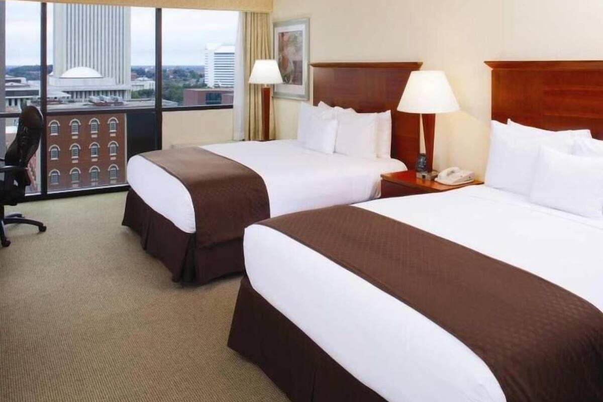 A hotel room with two beds, a desk, a chair, and a large window with a city view.