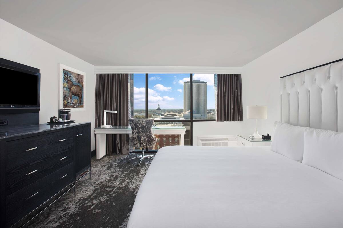 A hotel room with a large bed, a desk, a chair, a TV, and a view of a cityscape through the windows.