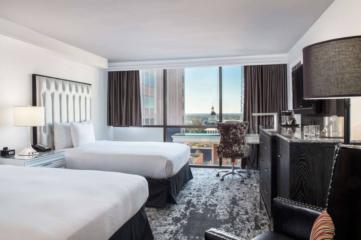 A hotel room with two beds, a desk with a lamp, a chair, and a view of a cityscape.