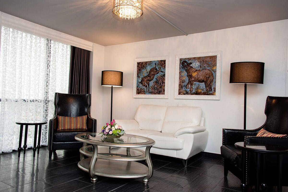 A modern living room with a white sofa, two black leather armchairs, a glass coffee table, and two wall-mounted lamps. There are two framed artworks depicting elephants.