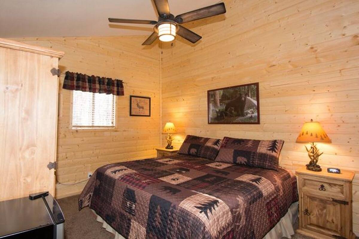 A wooden cabin room with a bed, two bedside lamps, a ceiling fan, a window with blinds, and a framed picture on the wall.