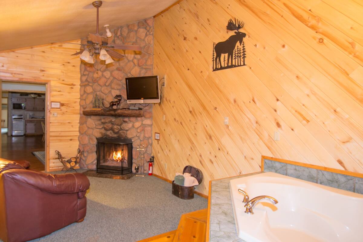 A log cabin interior with a fireplace, a television mounted above it, a leather sofa, a bathtub, and a moose wall decal.