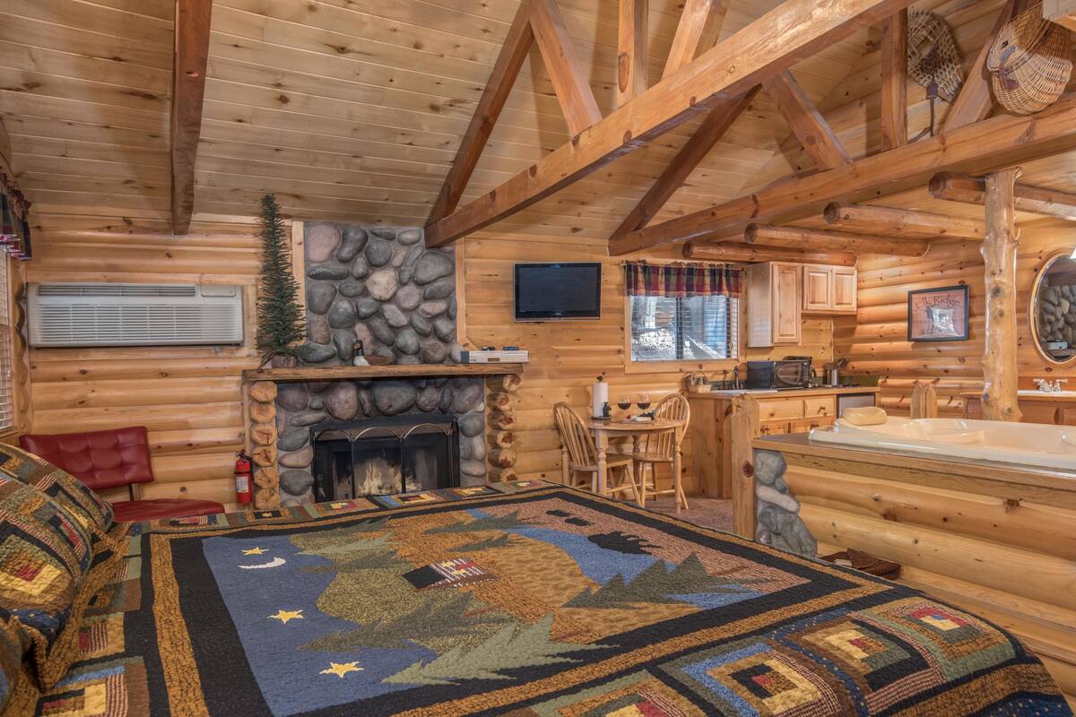 A log cabin interior with a stone fireplace, a dining area, a kitchen, and a bathroom with a large tub.