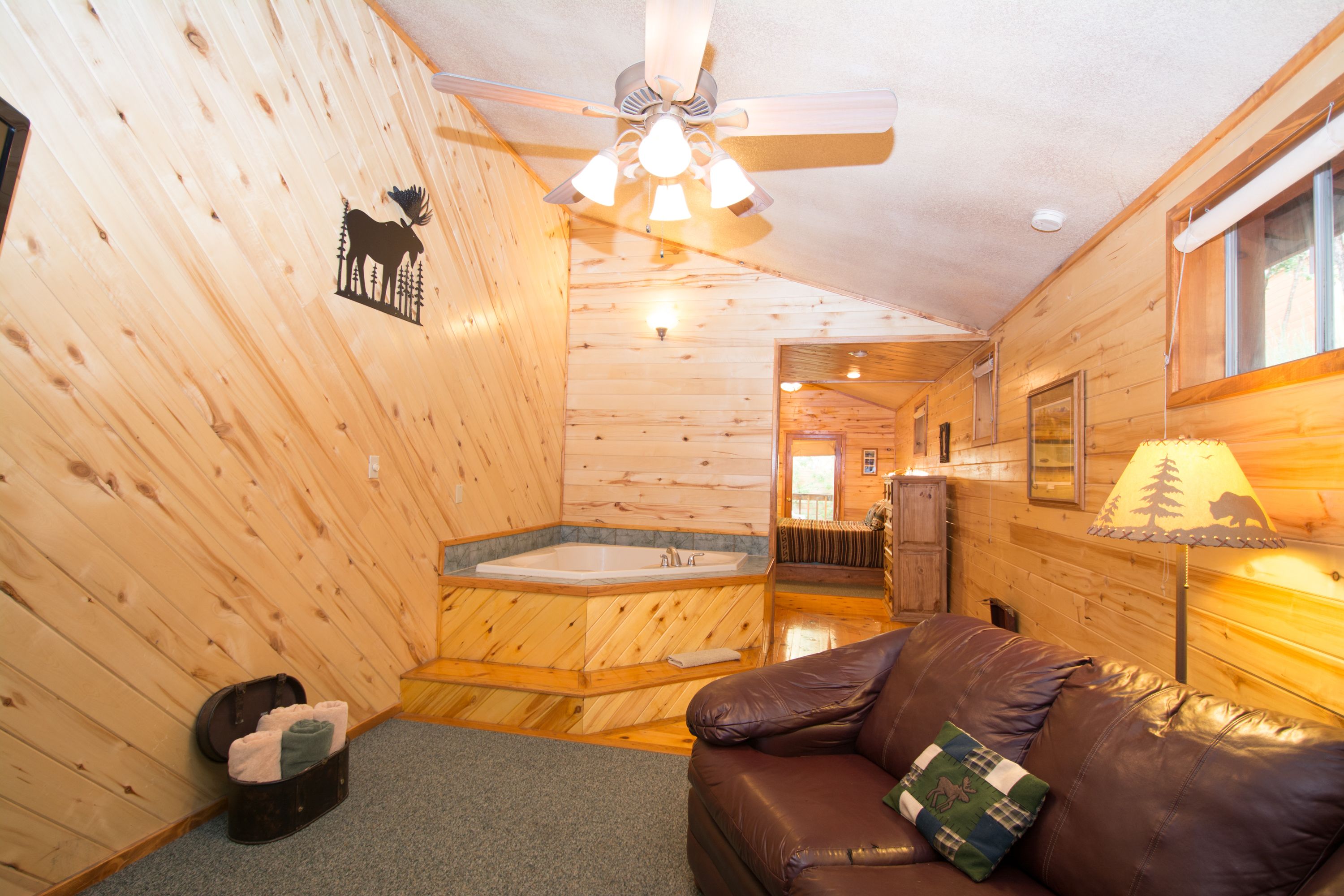 A cabin interior with a wooden interior, a brown leather sofa, a ceiling fan, a bathtub, and a lamp.