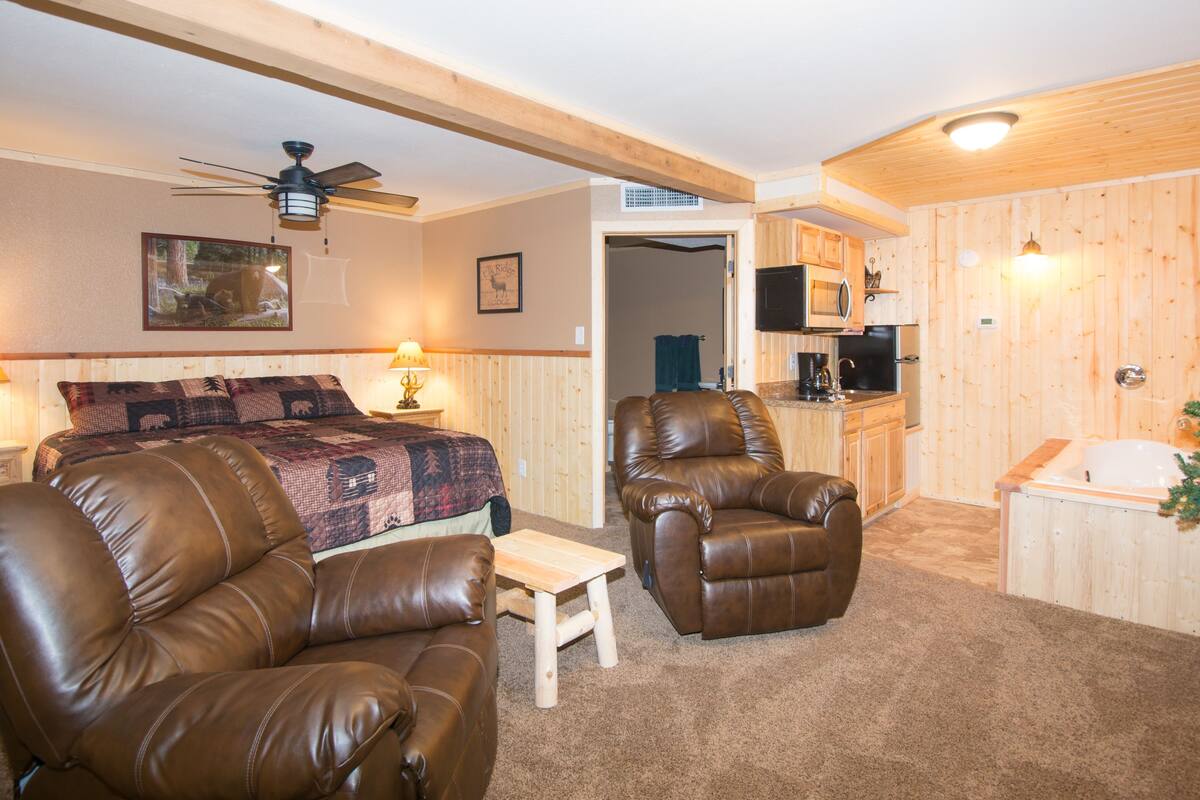 A cozy room with a bed, two leather chairs, a small table, a ceiling fan, and a kitchen area with wooden walls.
