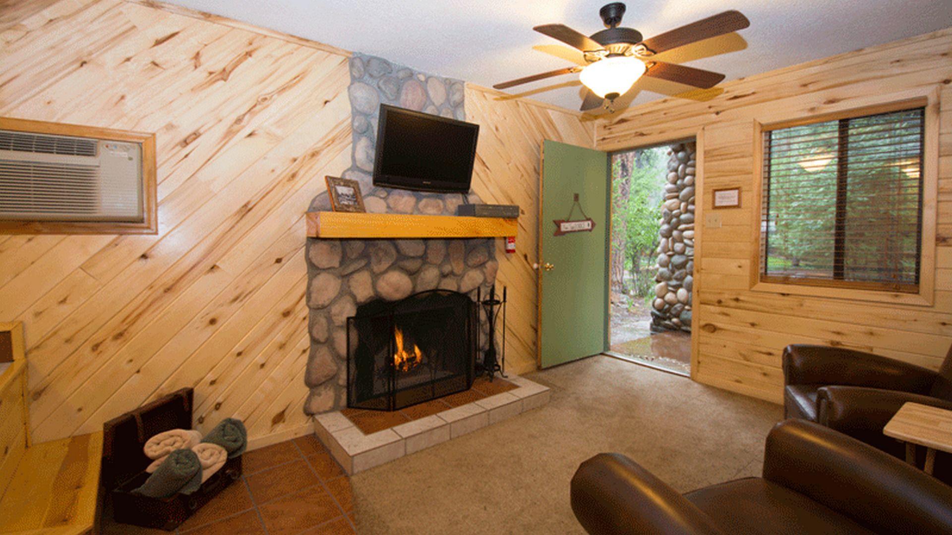 A cozy cabin interior with a stone fireplace, a television mounted above it, a ceiling fan, and a view through a door to an outdoor stone wall.
