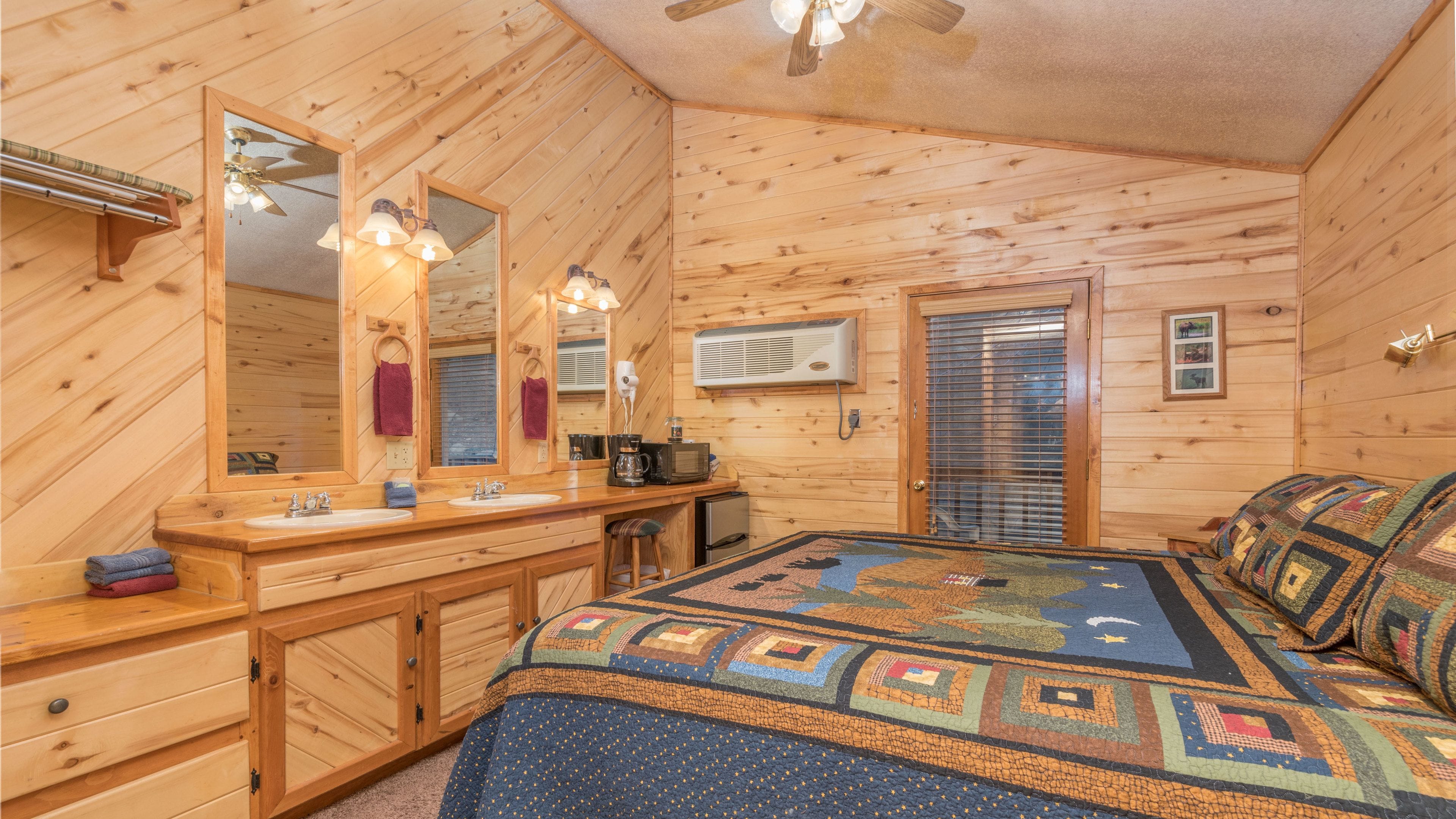 A wooden cabin bedroom with a large bed, a wooden vanity with a mirror, a ceiling fan, and a wall-mounted heater.