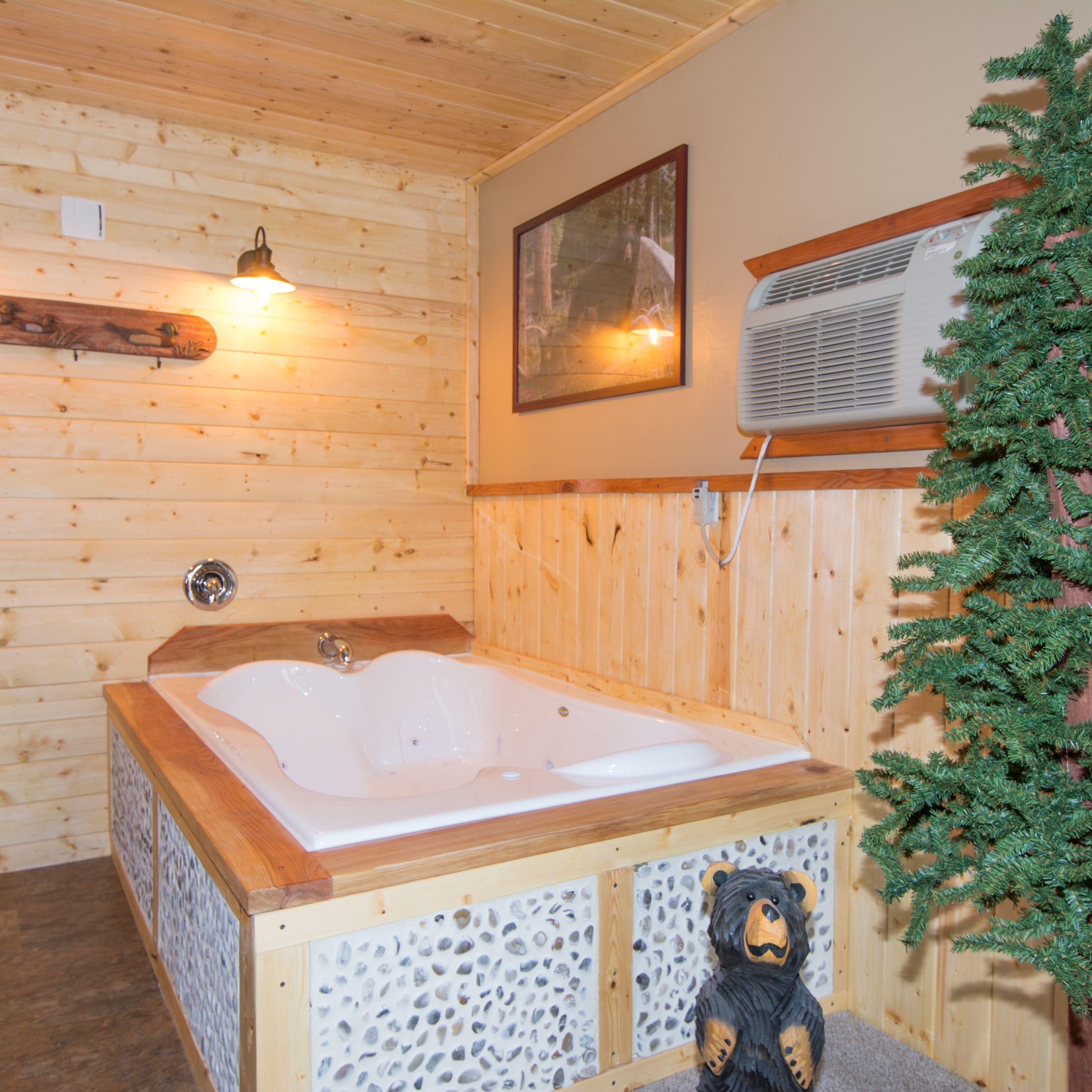 A wooden cabin interior with a wooden bathtub, a bear decoration, a wall-mounted air conditioner, and a framed picture.
