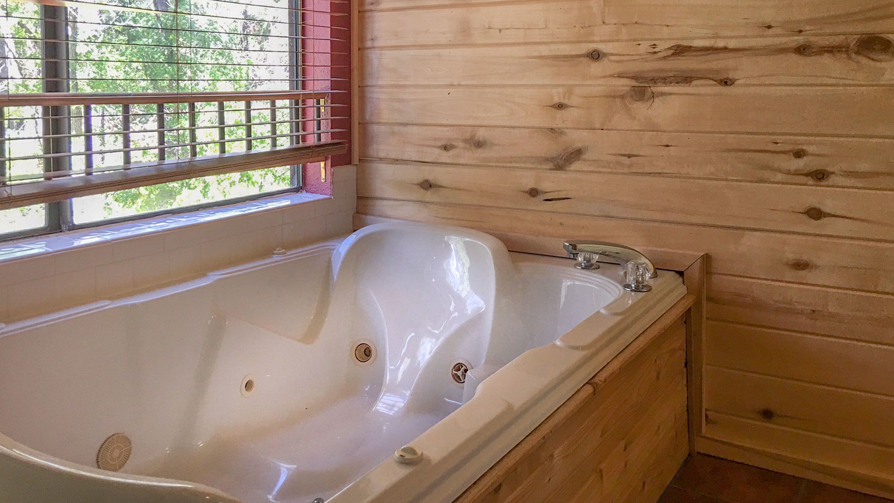 A wooden cabin with a large bathtub, a window with blinds, and a view of greenery outside.