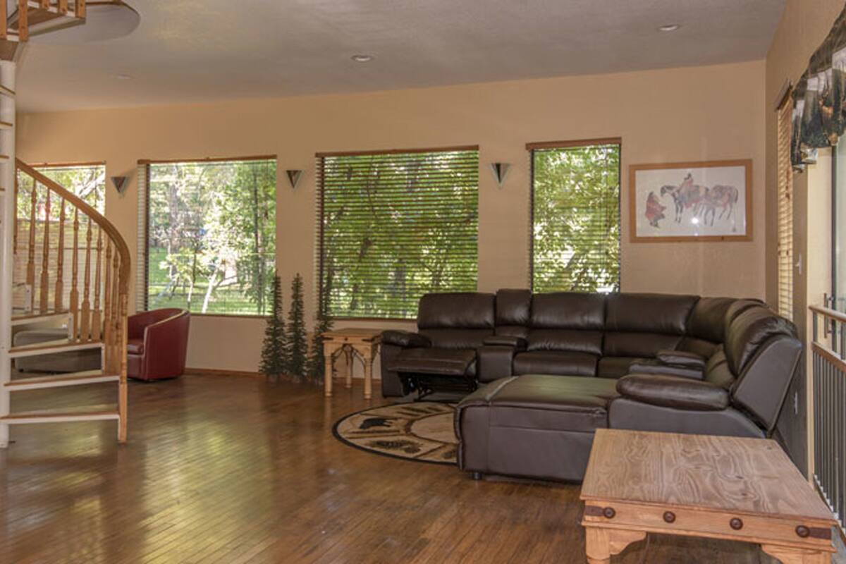 A spacious living room with a wooden staircase, a leather sofa, and large windows with blinds.