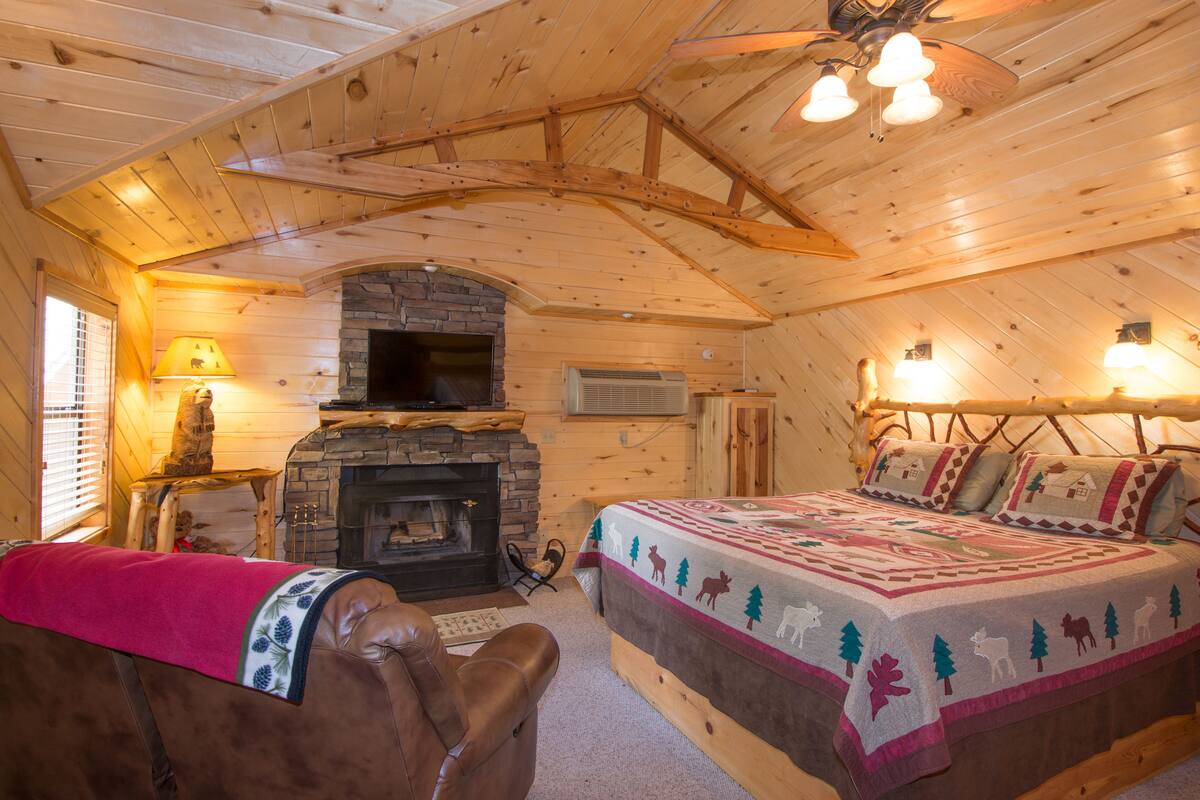 A cozy log cabin room with a fireplace, a bed with a quilt, a leather chair, and a ceiling fan.