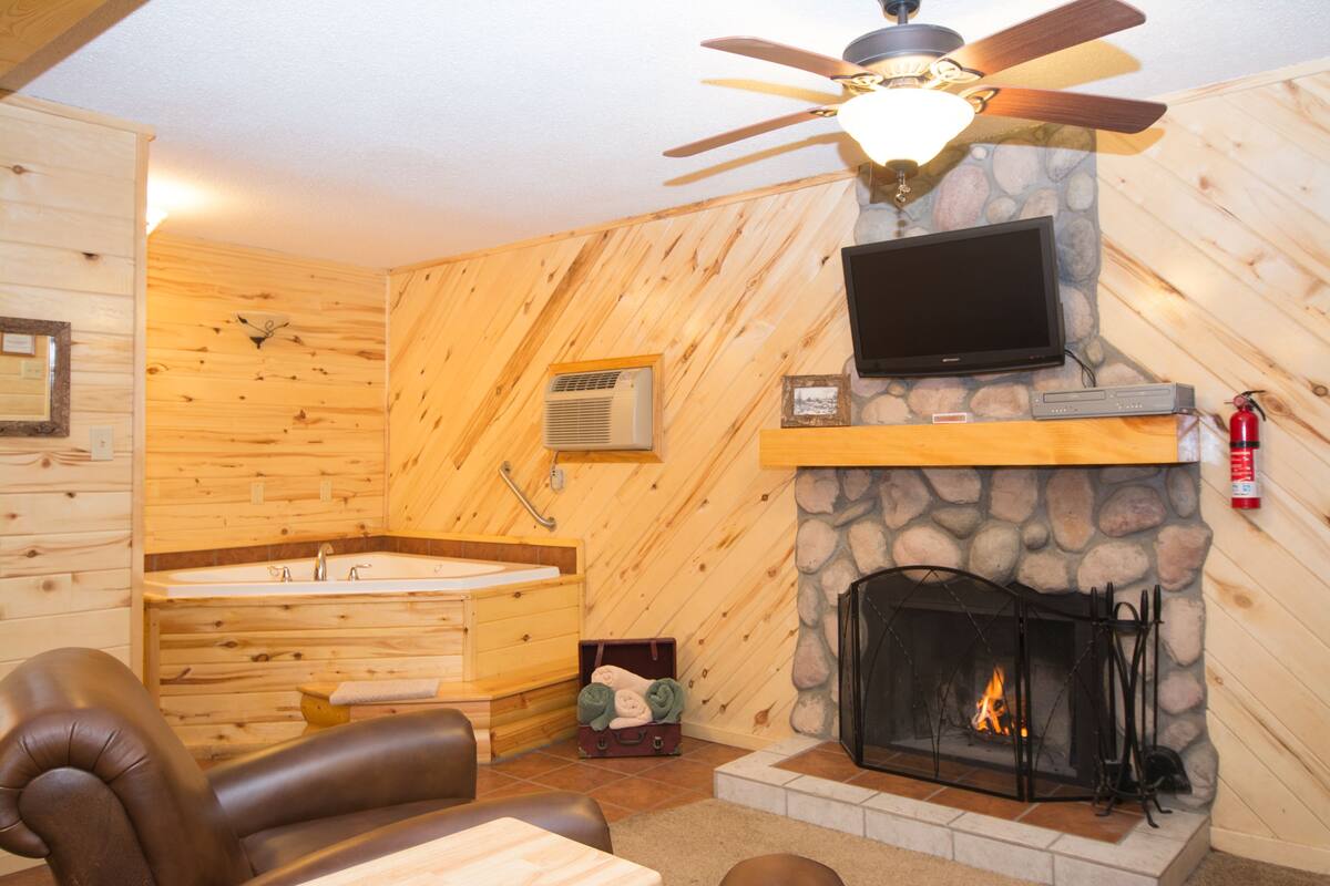 A log cabin interior with a stone fireplace, a television mounted above it, a ceiling fan, and a wooden wall with a built-in bathtub.