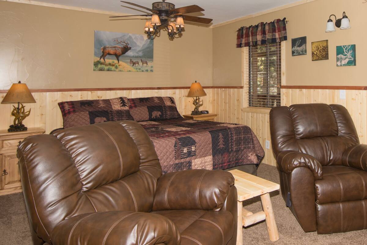 A room with a bed, two leather chairs, a wooden table, a ceiling fan, and wall art.