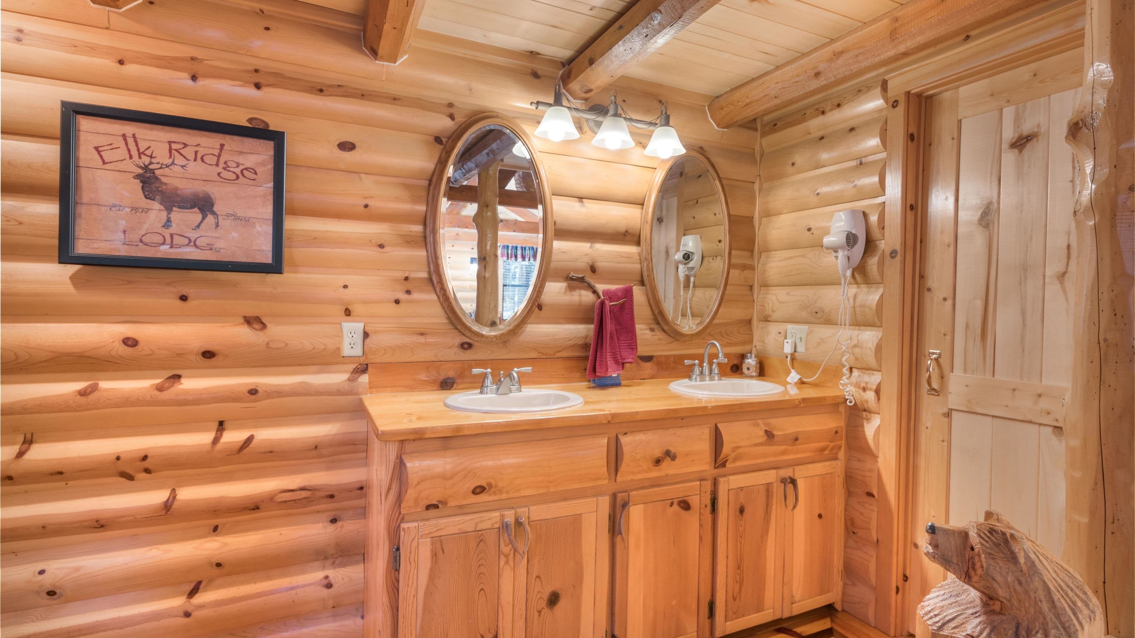 A wooden cabin bathroom with two sinks, a mirror, and a framed picture of an elk.