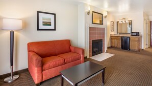A hotel room with a red sofa, a small table, a floor lamp, a fireplace, a kitchen area with a microwave, and a framed picture on the wall.