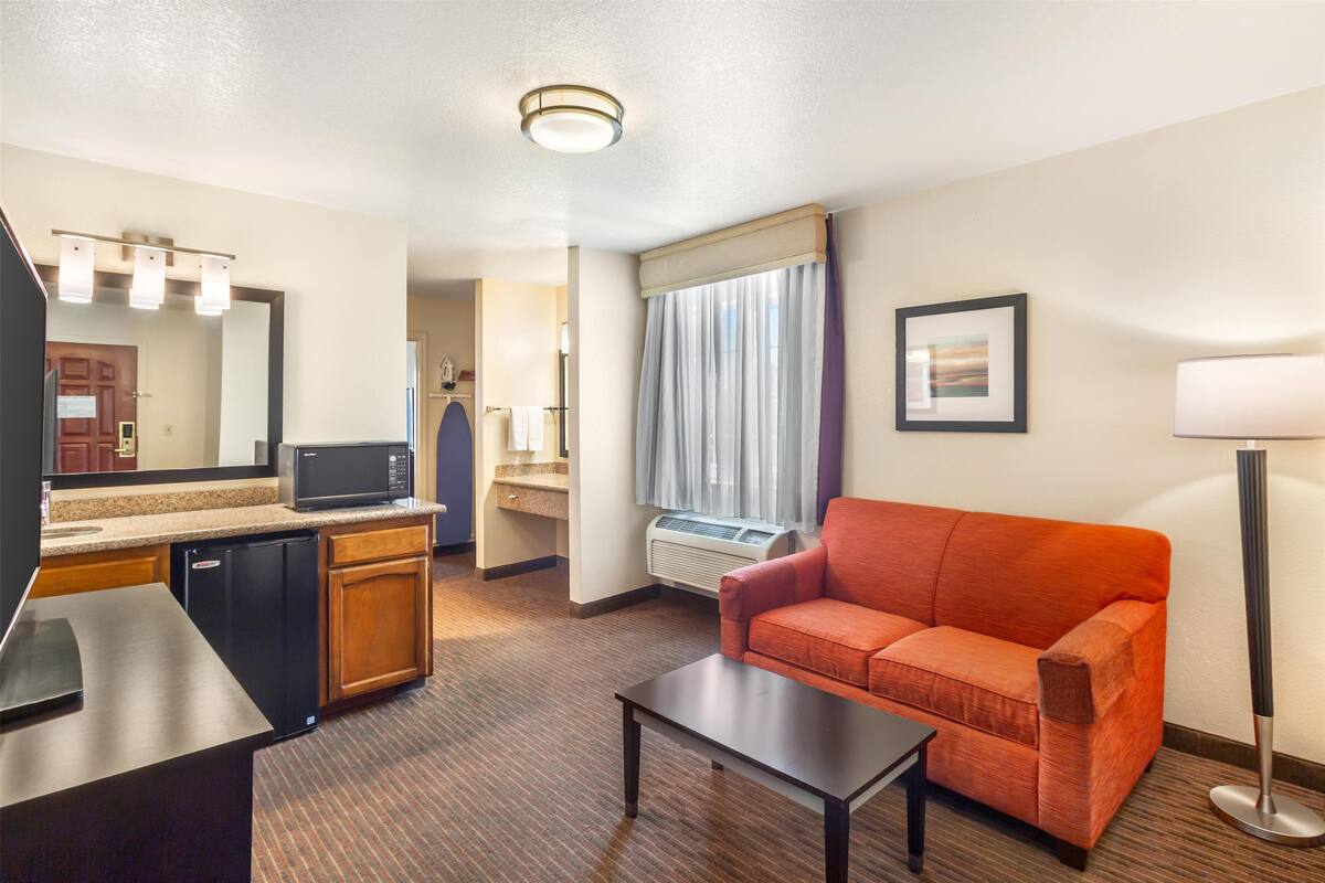 A hotel room with a kitchenette, a sofa, a coffee table, a flat-screen TV, a mirror, a window with curtains, and a floor lamp.