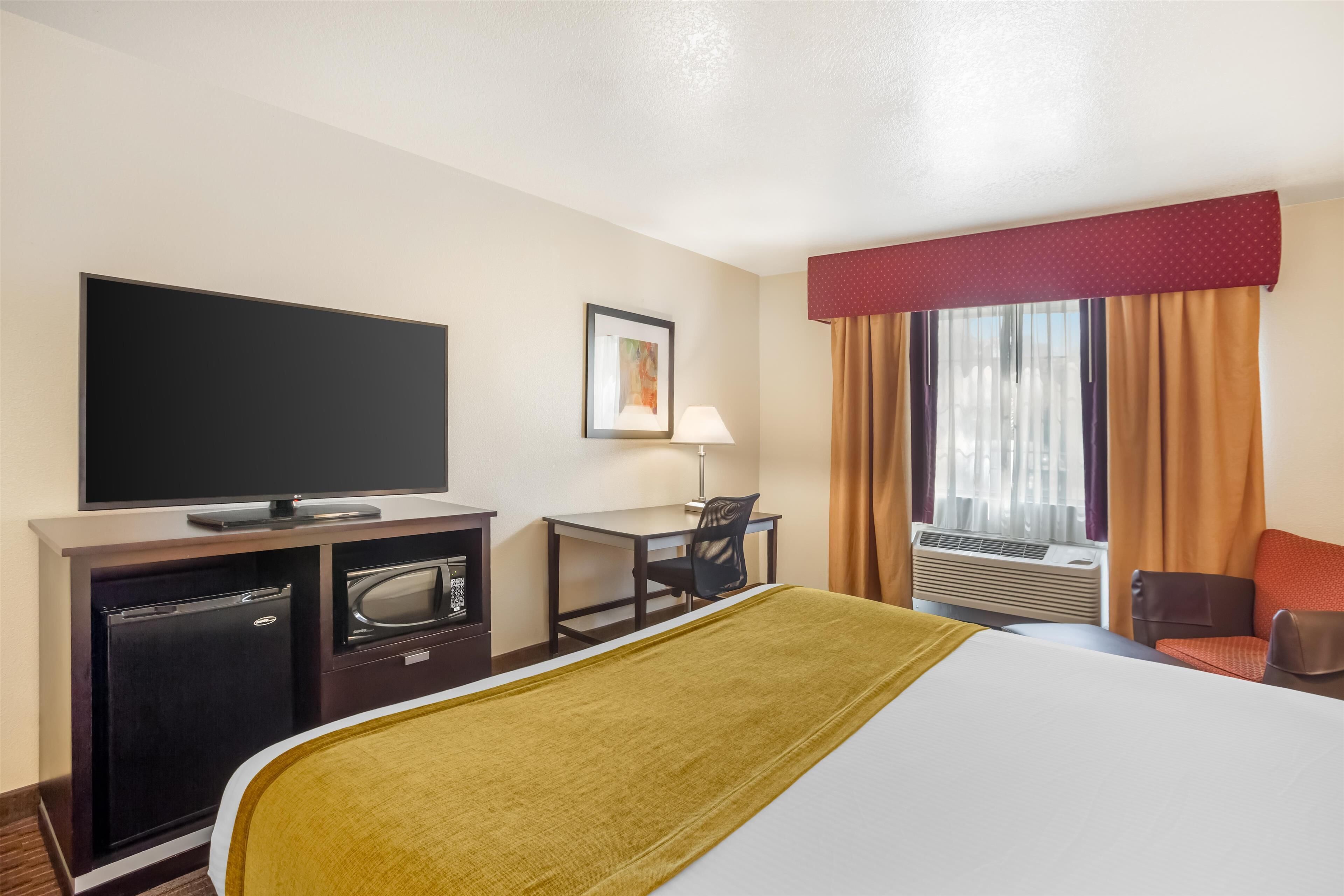Habitació d'hotel amb llit, escriptori, televisió, nevera petita i finestra amb cortines.