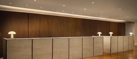 Un mostrador de recepción con un panel de madera de fondo, luces empotradas en el techo y un suelo de madera pulida.