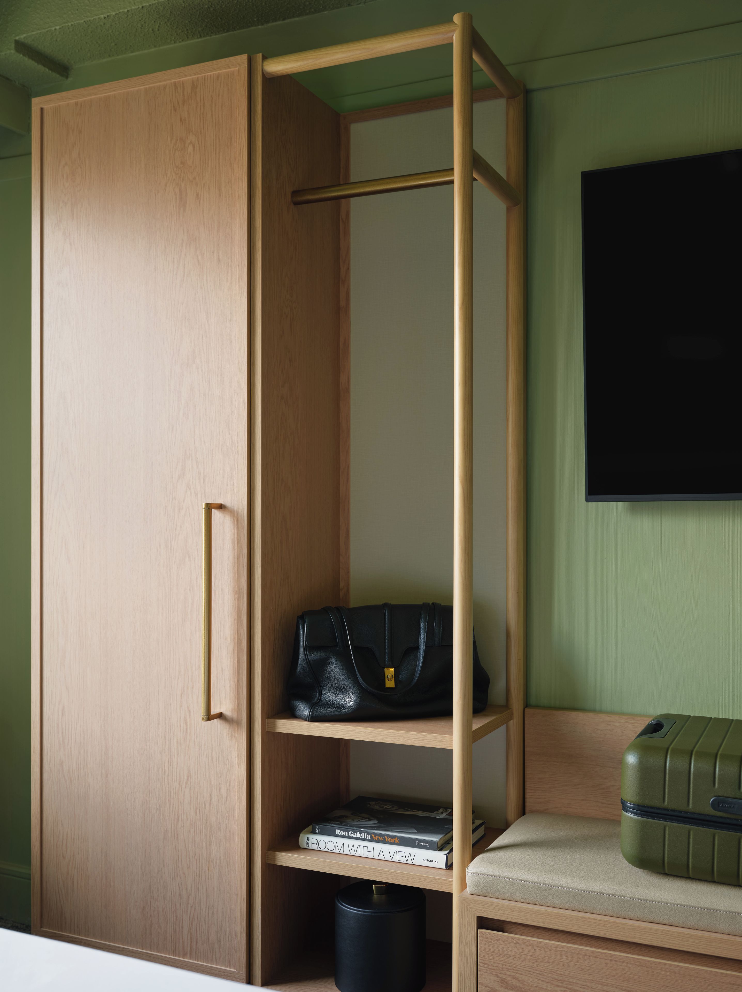 Una habitación con un armario de madera, una estantería con un bolso negro y libros, una maleta verde, y un televisor en la pared.