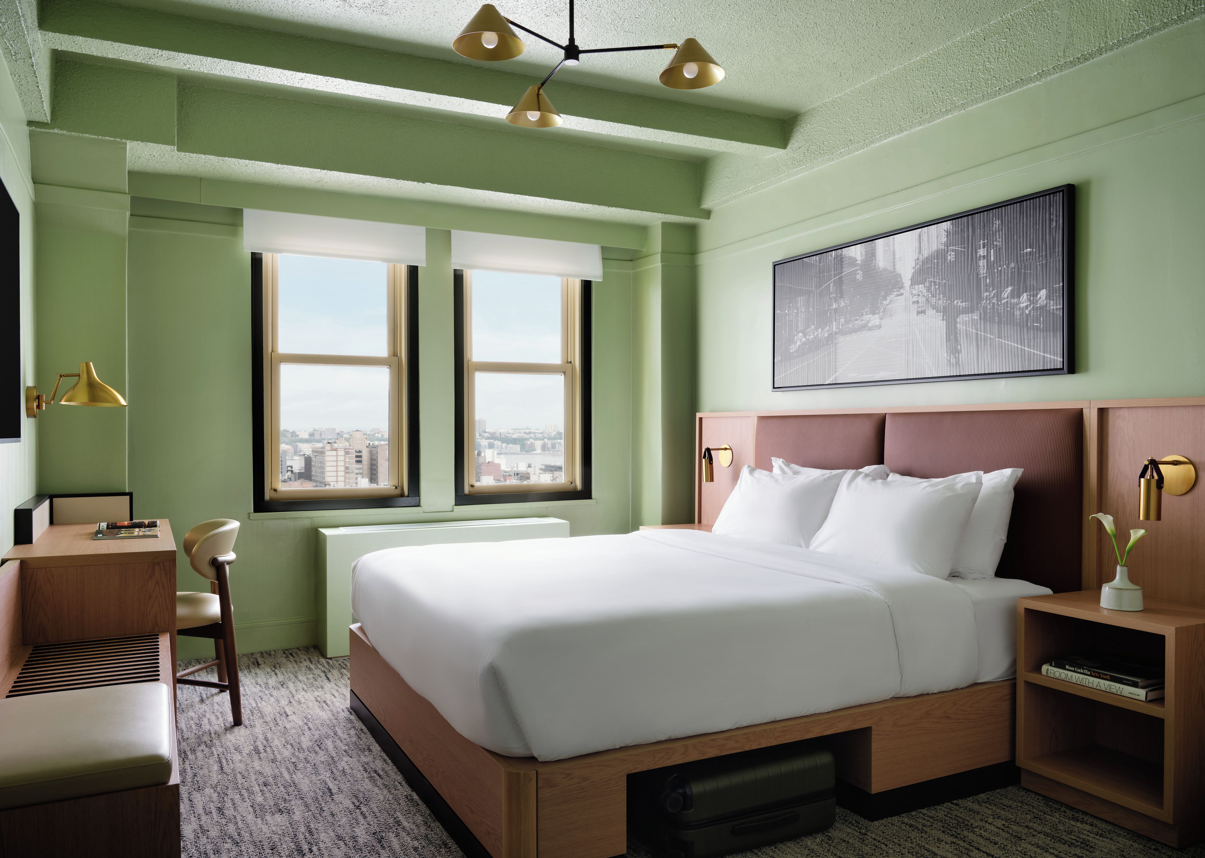 A hotel room with a large bed, a desk, a chair, a radiator, a window with a view of the city, and a painting on the wall.