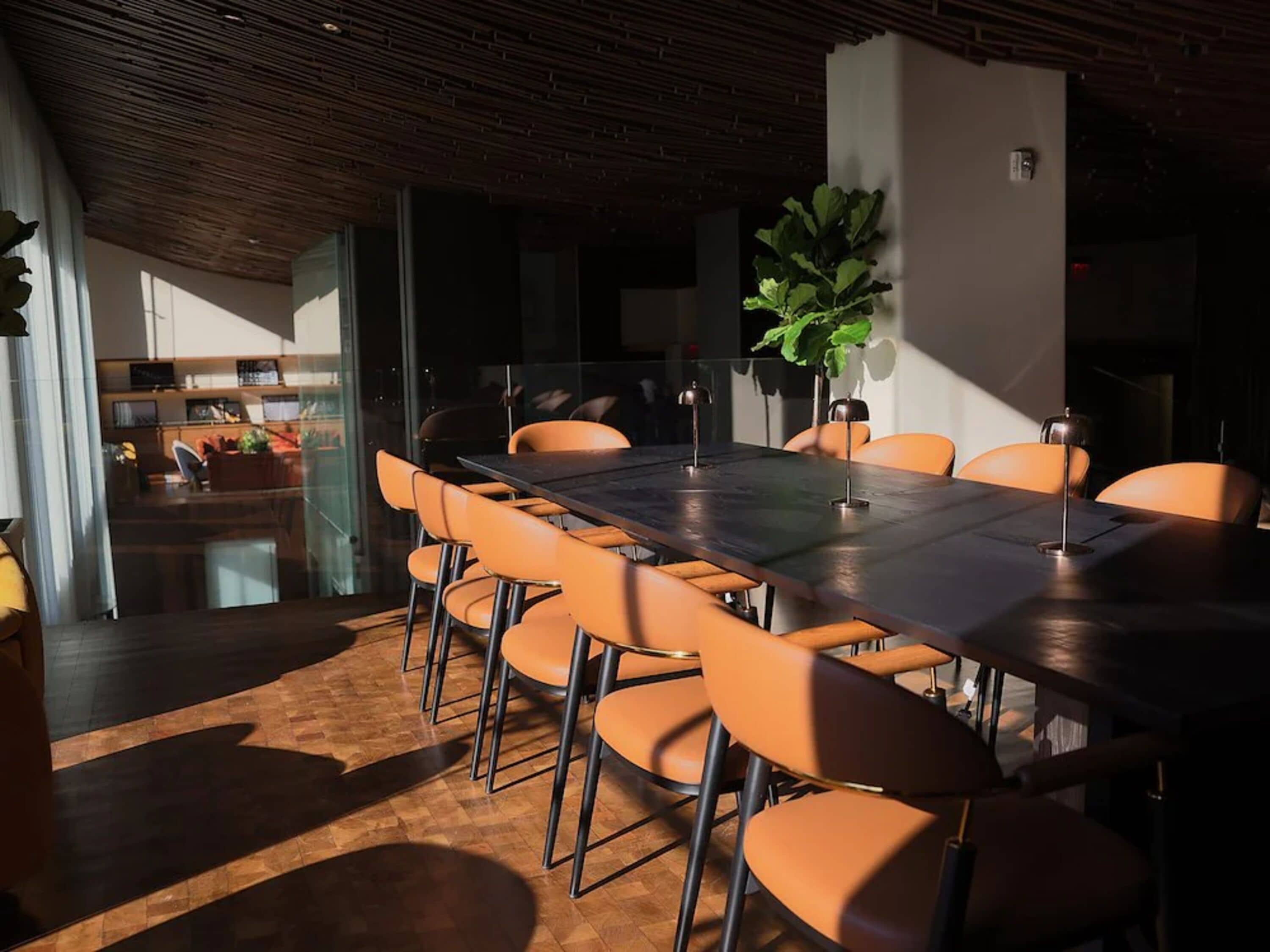 A modern dining area with a long table, orange chairs, and a potted plant.