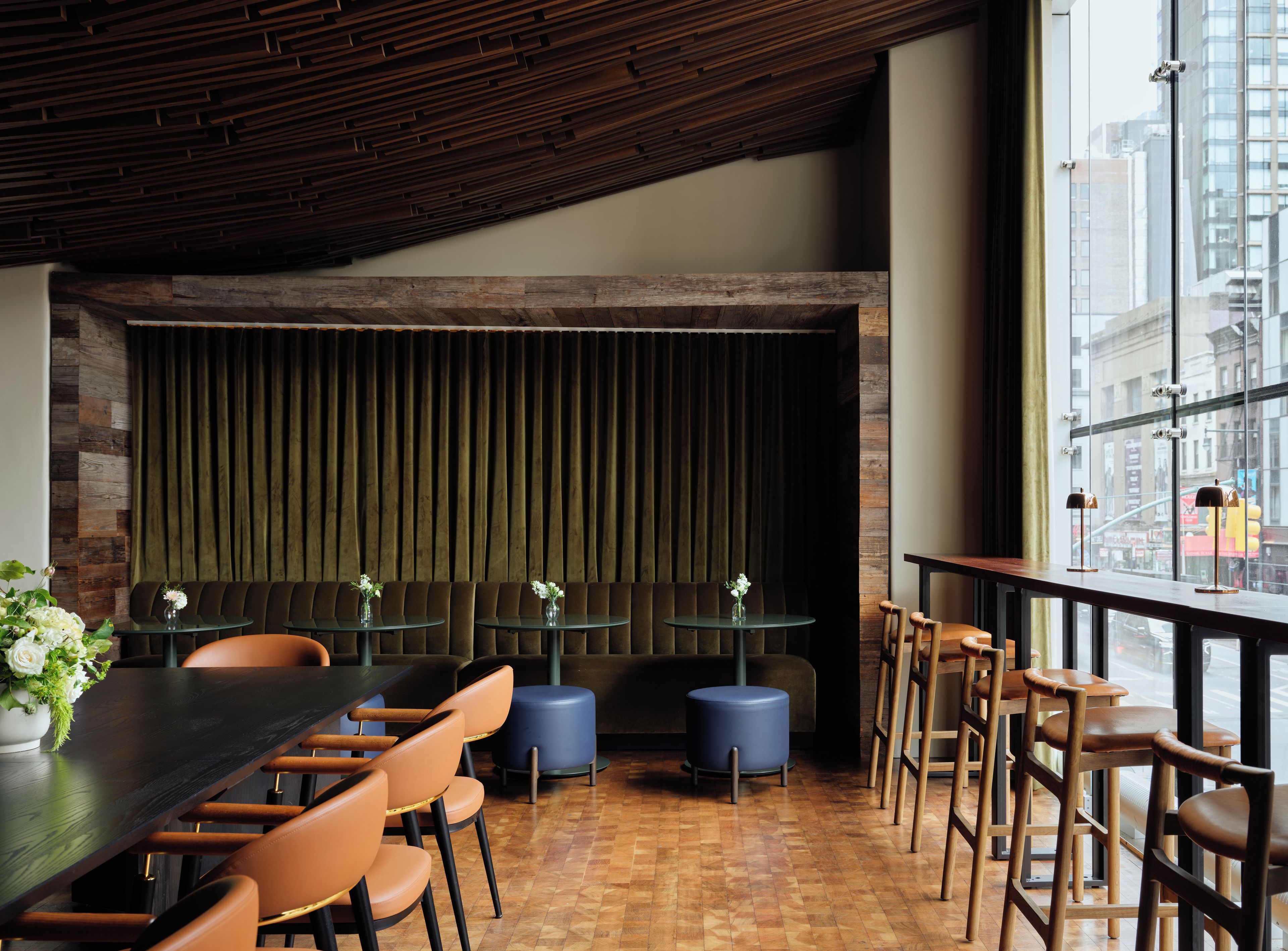 A restaurant interior with a wooden ceiling, dark green upholstered booth seating, round tables, and a view of the city through large windows.