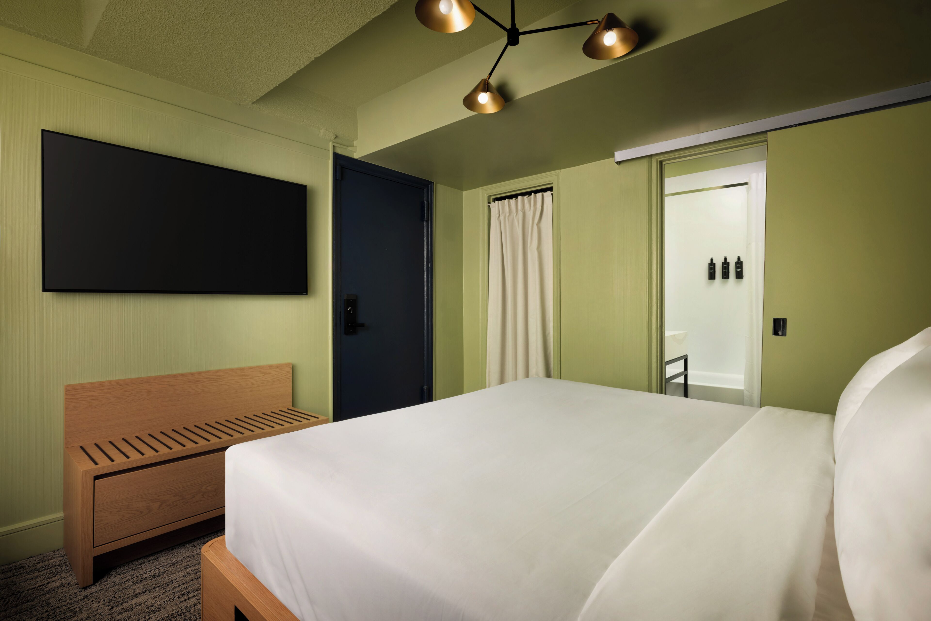 A hotel room with a bed, a wooden headboard, a flat-screen TV mounted on the wall, a door, and a ceiling light fixture.