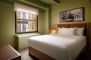 Un dormitorio de hotel con una cama grande, una ventana que ofrece vistas a edificios, una mesita de noche con lámpara y un cuadro enmarcado en la pared.