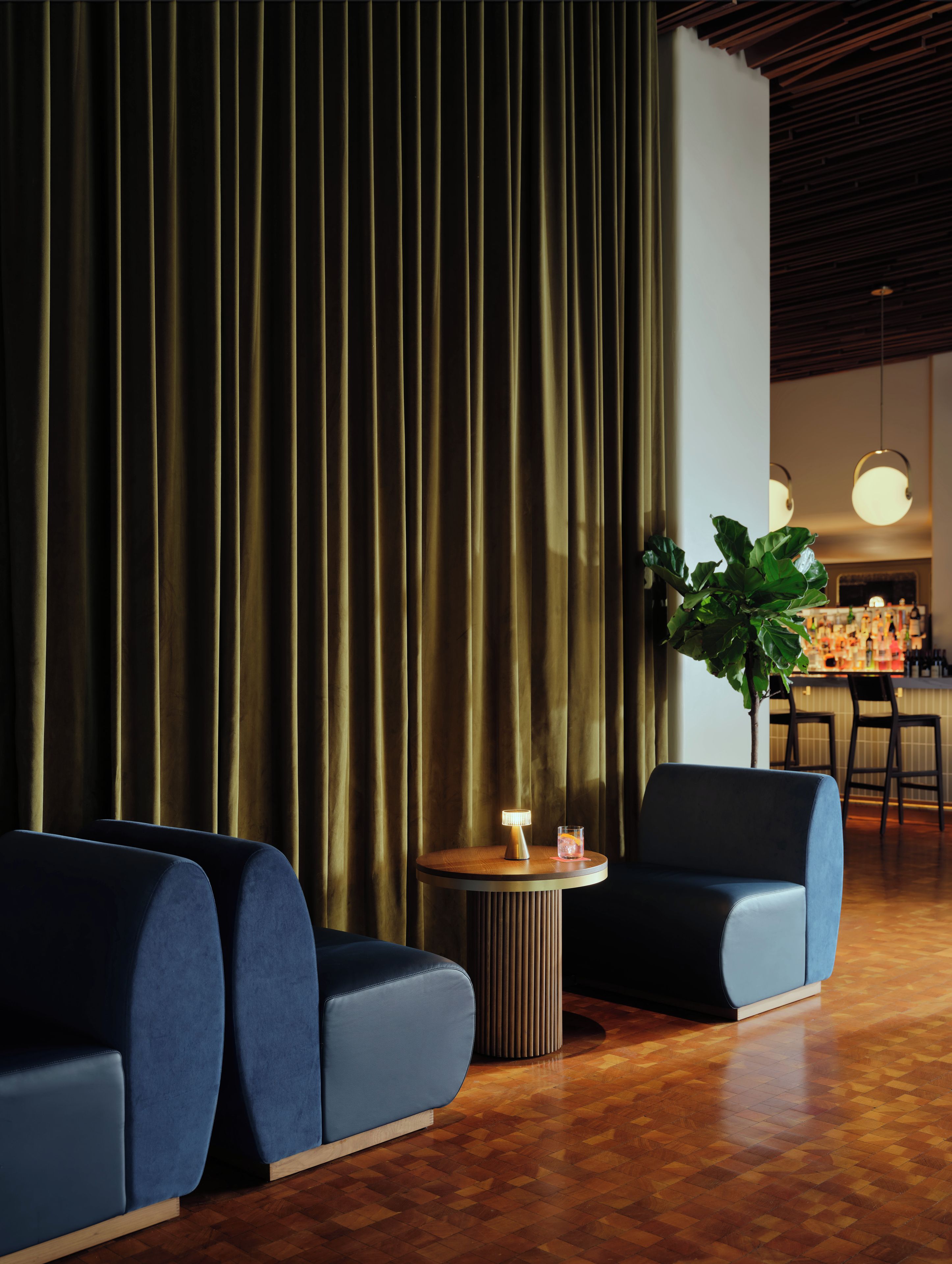 Un interior moderno con una cortina verde oscuro, una mesa redonda de madera con una lámpara y dos sofás azules.