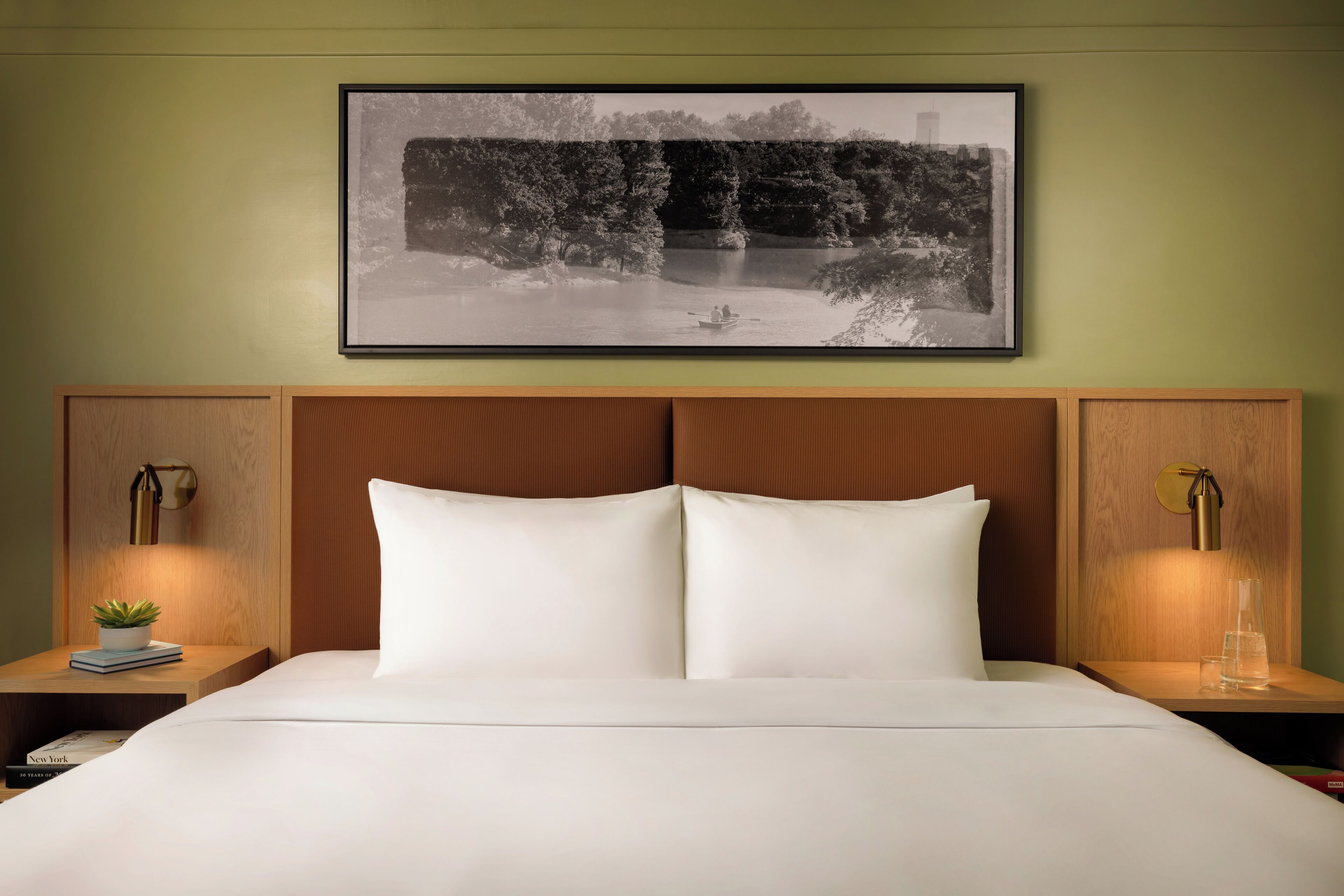 Una cama con sábanas blancas, cabecera con paneles de madera, dos mesitas de noche con lámparas y una fotografía en blanco y negro enmarcada sobre la cama.