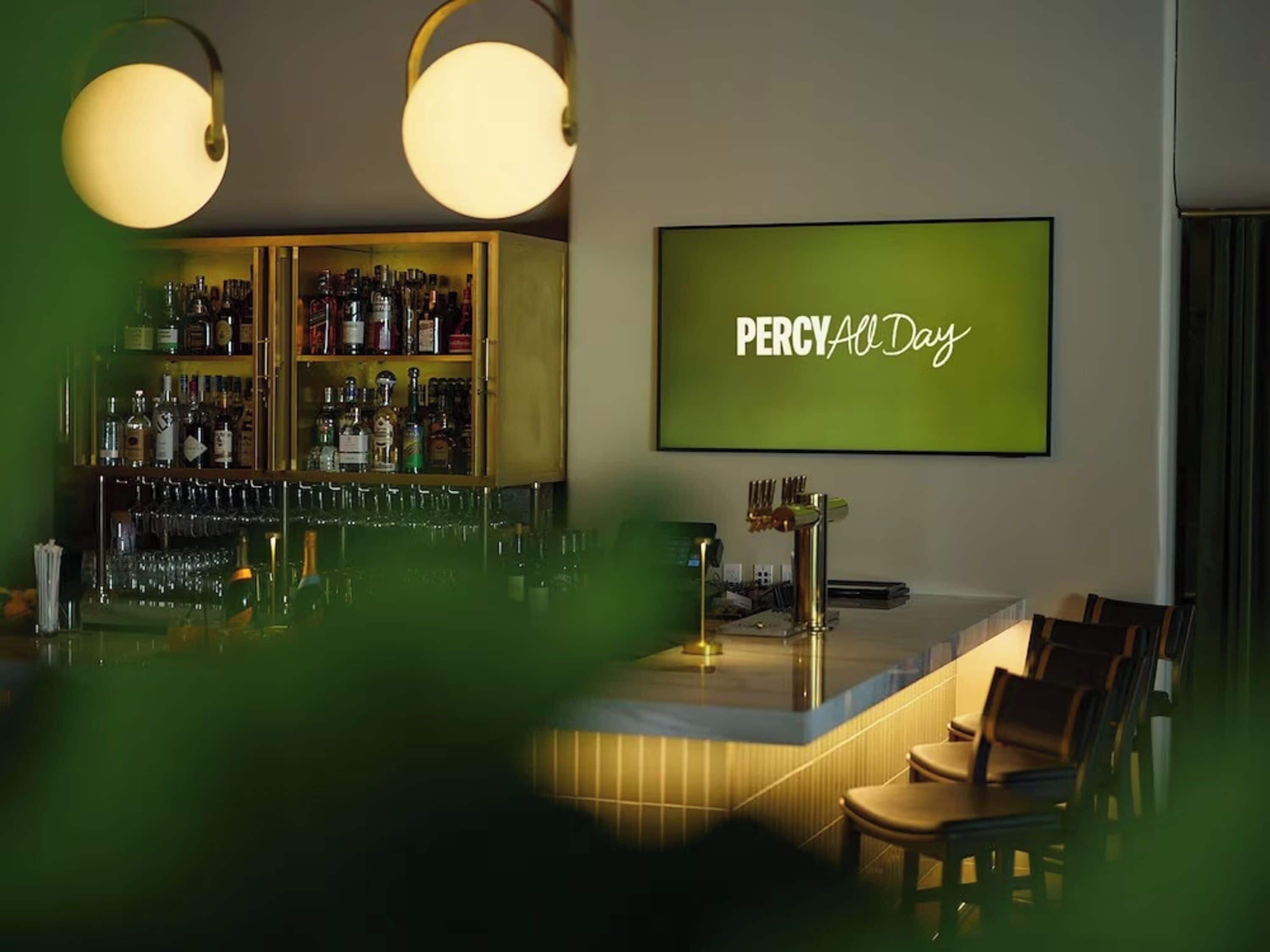 A bar area with a neon sign reading 'PERCY All Day' and a shelf of liquor bottles.
