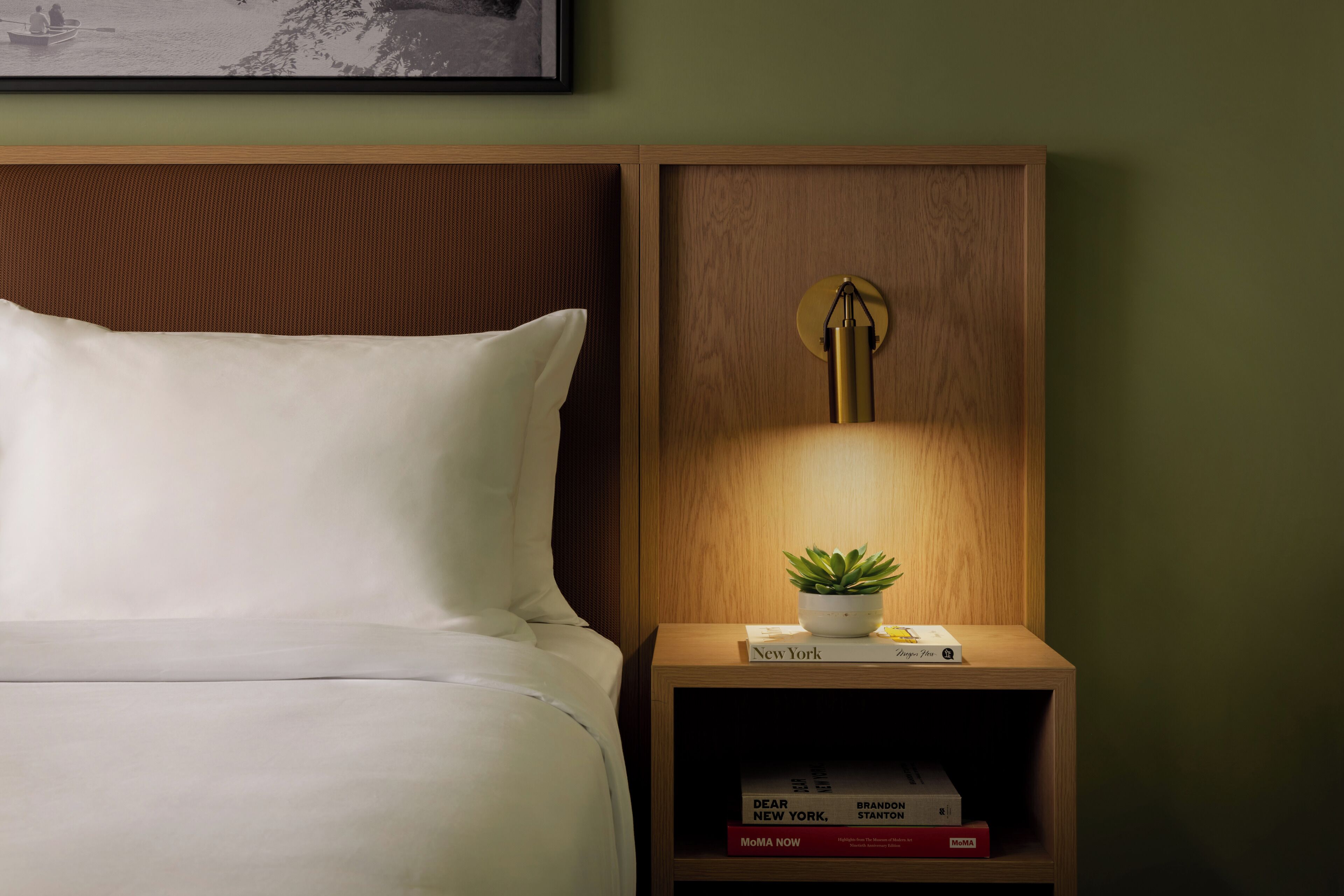 A neatly made bed with a pillow, a bedside table with a lamp, a small plant, and a few books.