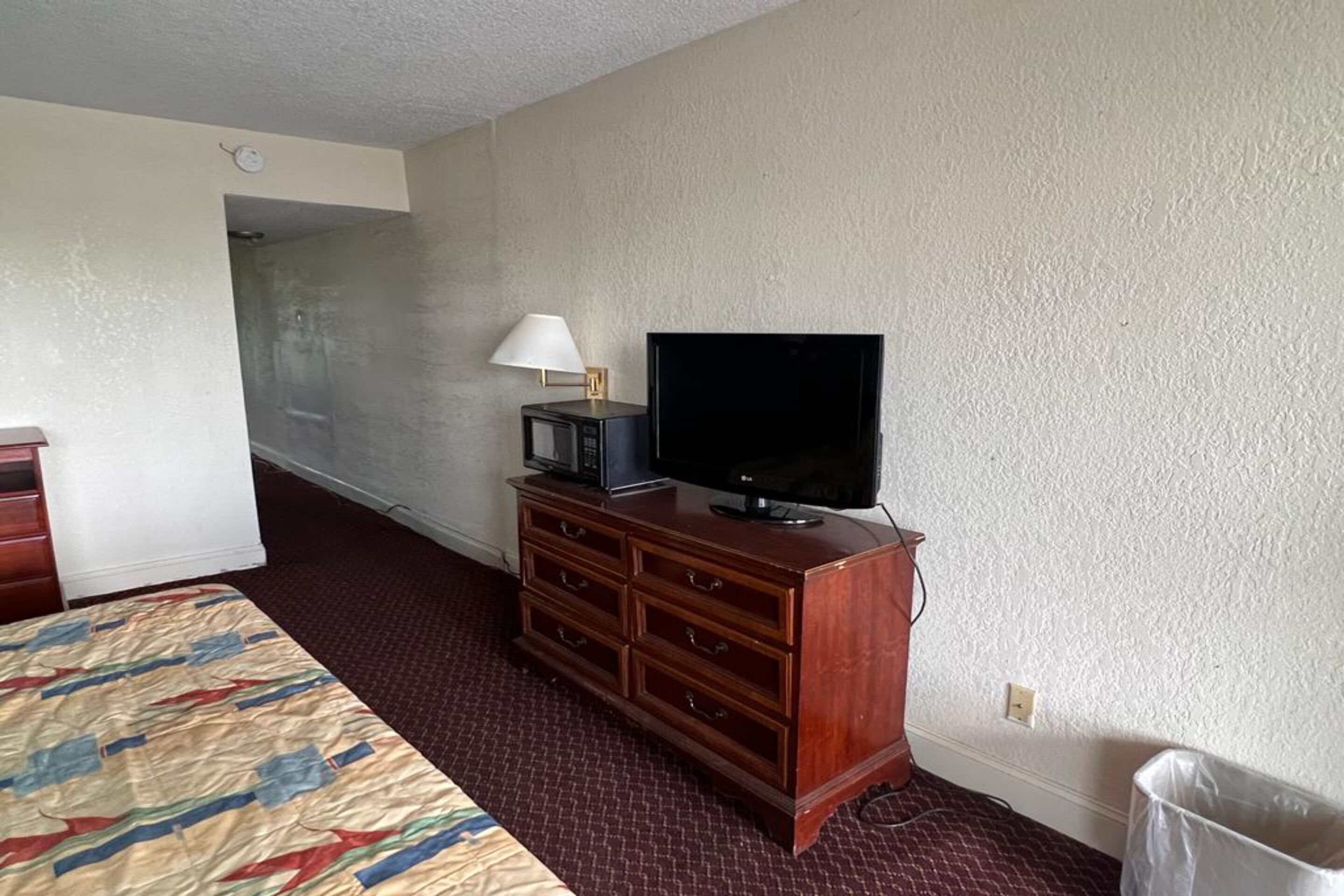 A hotel room with a bed, a wooden dresser, a television, and a lamp.