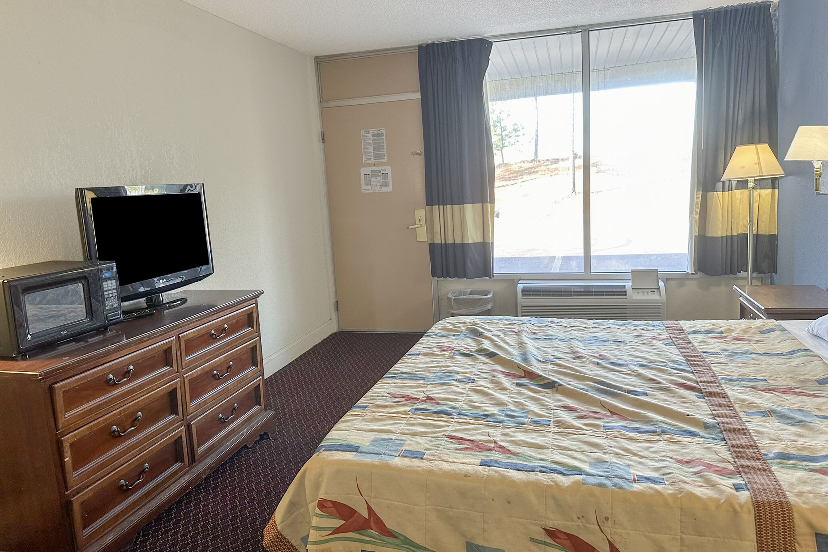 A hotel room with a bed, a dresser with a television, a window with curtains, and a lamp.
