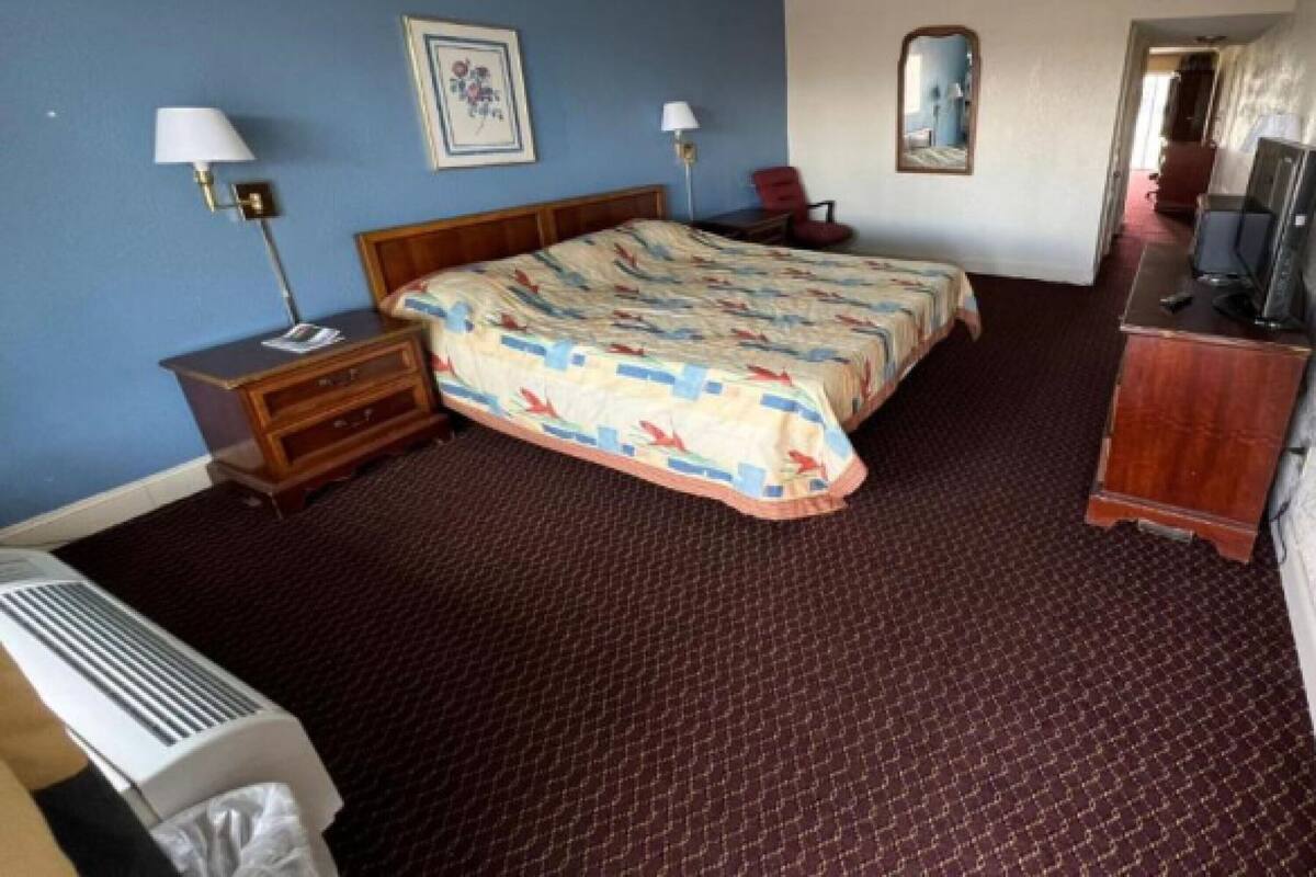 Ein Hotelzimmer mit einem großen Bett, zwei Nachttischen mit Lampen, einem Schreibtisch mit Computer, einem Fernseher und einem Spiegel an der Wand.