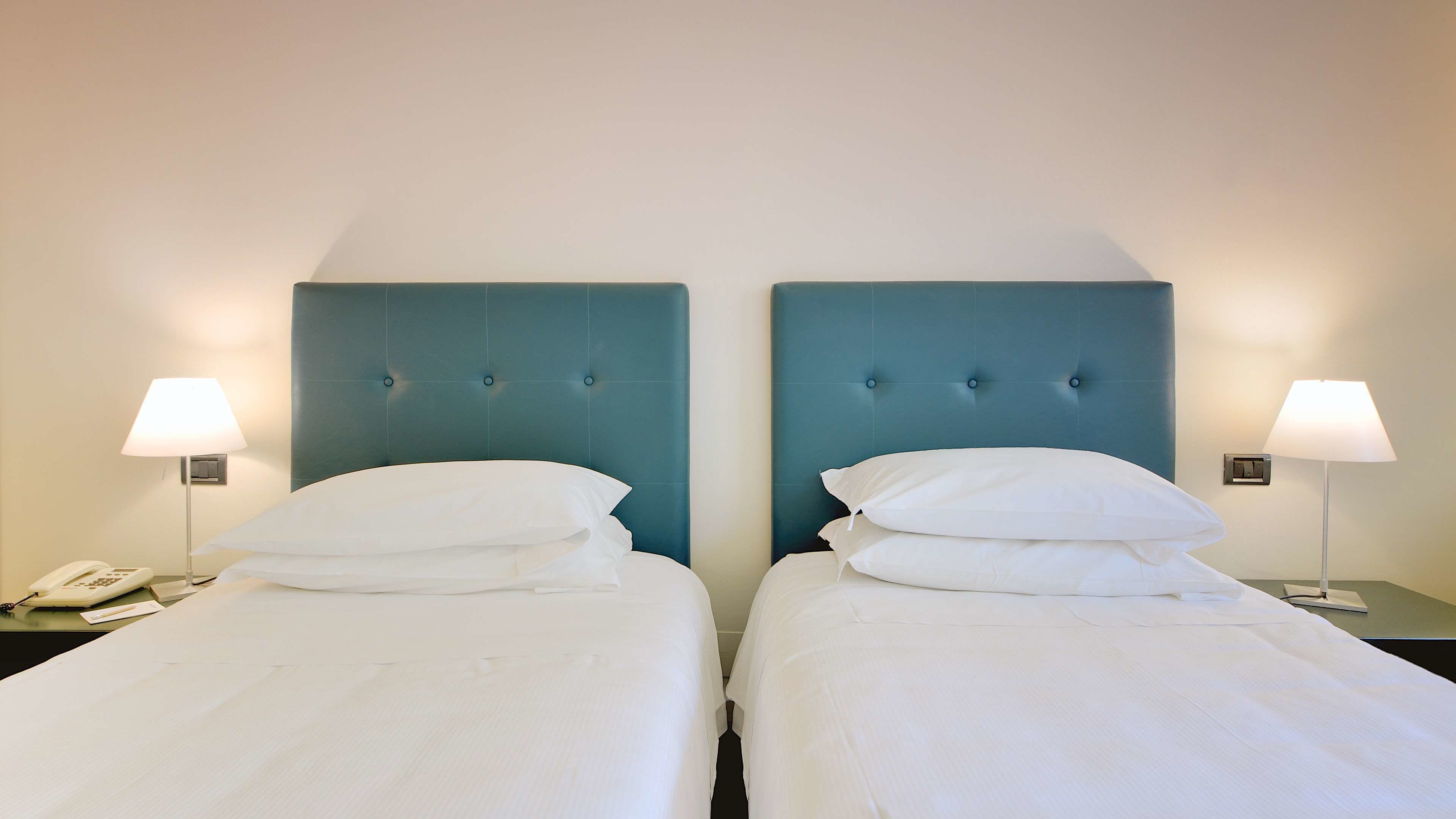 Twee eenpersoonsbedden met witte lakens, een blauw hoofdbord, bedlampjes en een telefoon op het nachtkastje links.