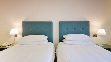 Twee eenpersoonsbedden met witte lakens, een blauw hoofdbord, bedlampjes en een telefoon op het nachtkastje links.