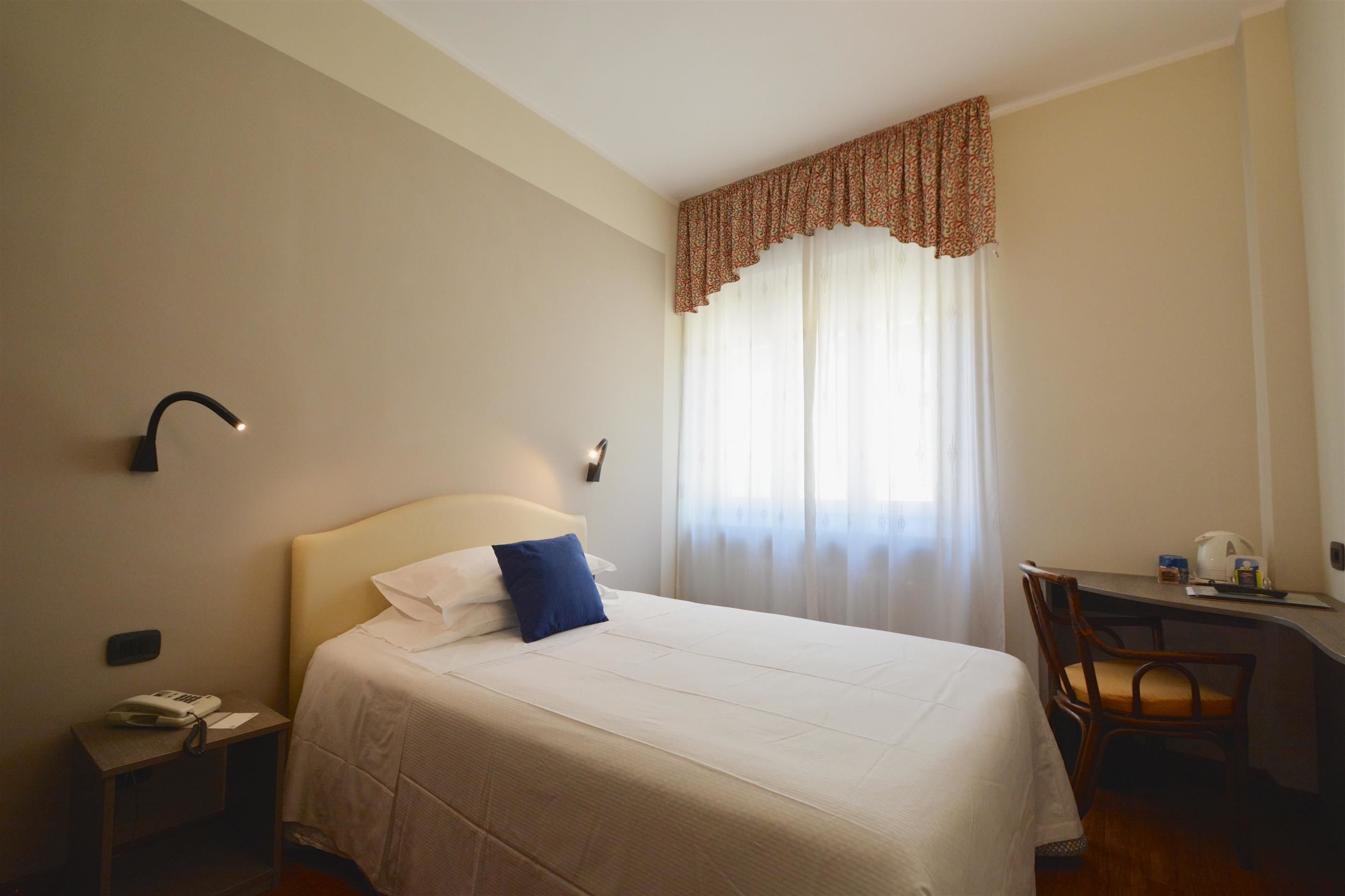 A hotel room with a bed, a desk, a chair, a telephone, and a window with curtains.