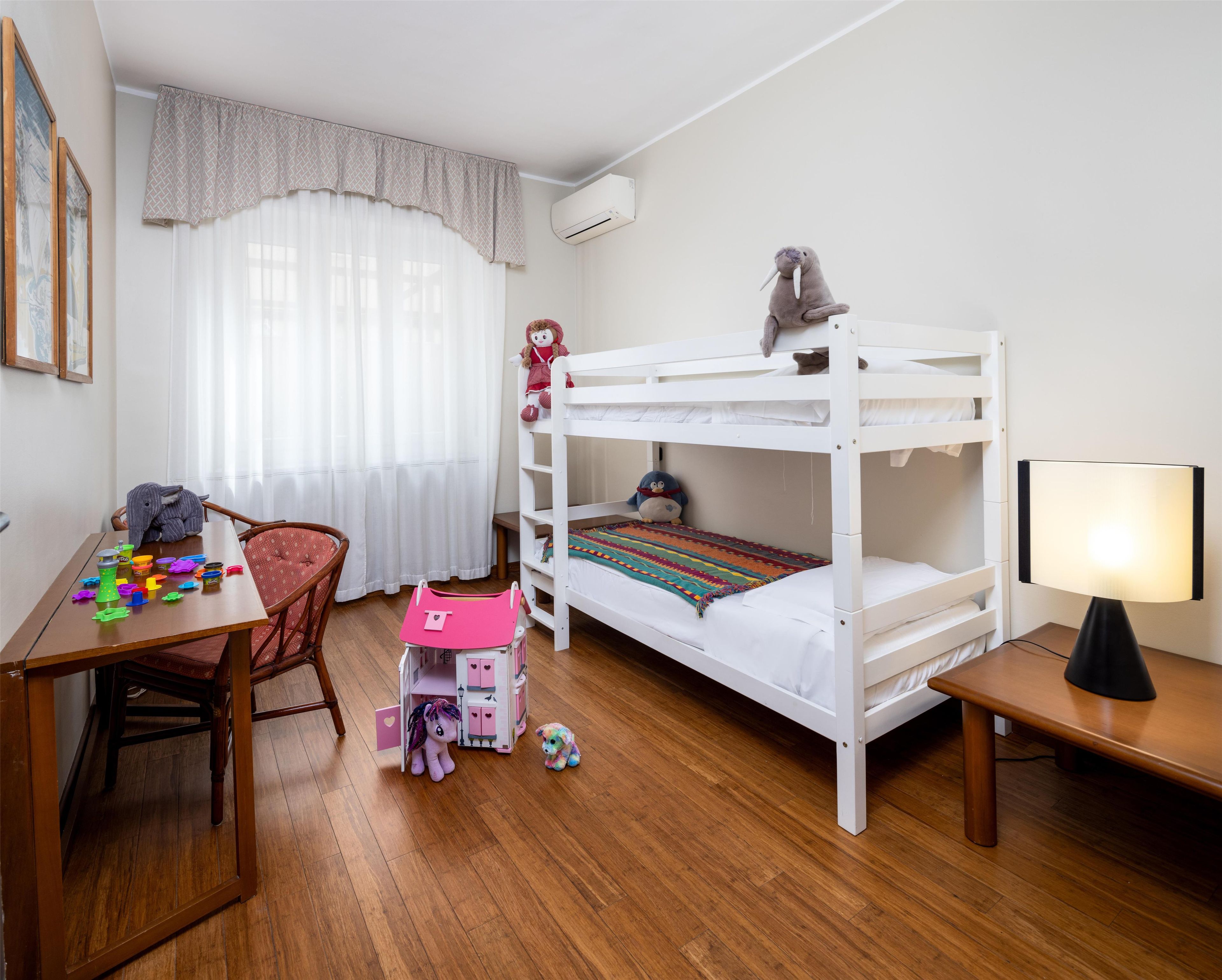 Uma cama beliche com um bicho de pelúcia em cima, uma escrivaninha com brinquedos, uma cadeira, um abajur e uma janela com cortinas.
