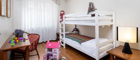 A bunk bed with a stuffed animal on top, a desk with toys, a chair, a lamp, and a window with curtains.