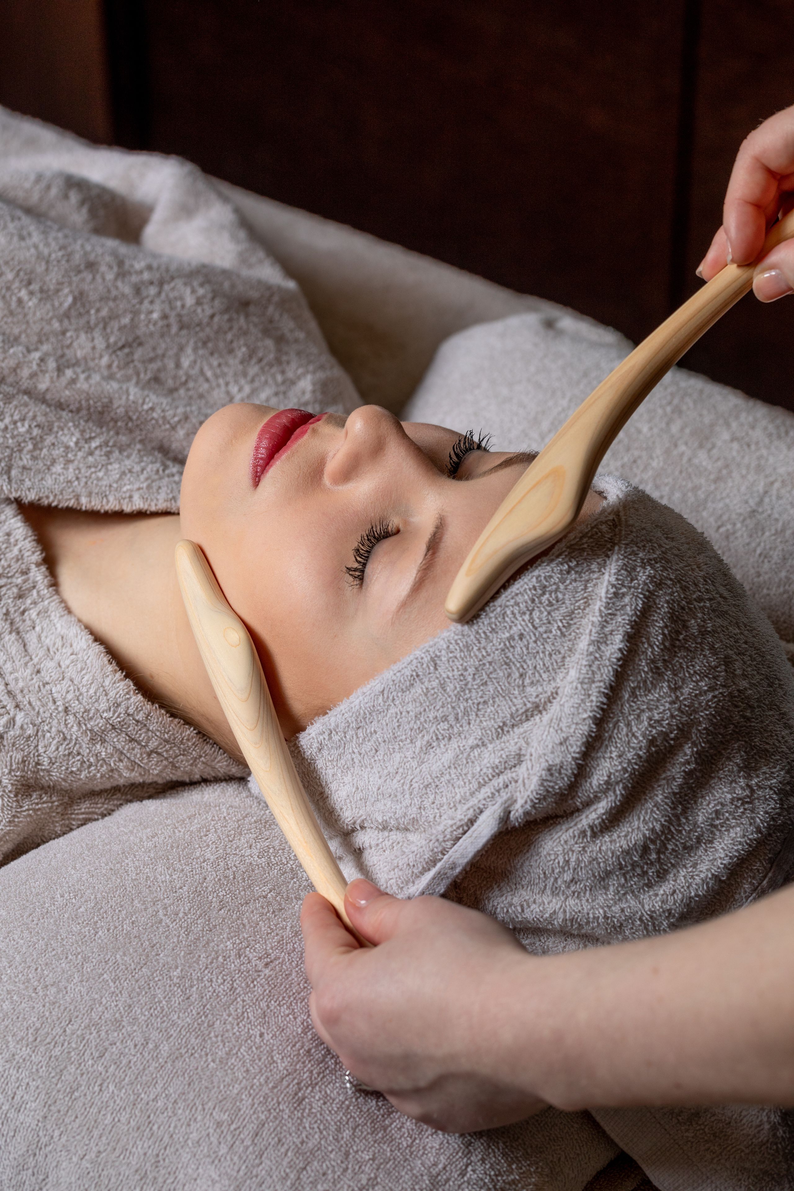 A person lying down with a towel wrapped around their head, receiving a facial treatment with wooden tools.