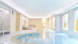 Una piscina interior de forma larga y estrecha, rodeada por un suelo y paredes con azulejos. Hay sillas de descanso y un banco a lo largo de la piscina.