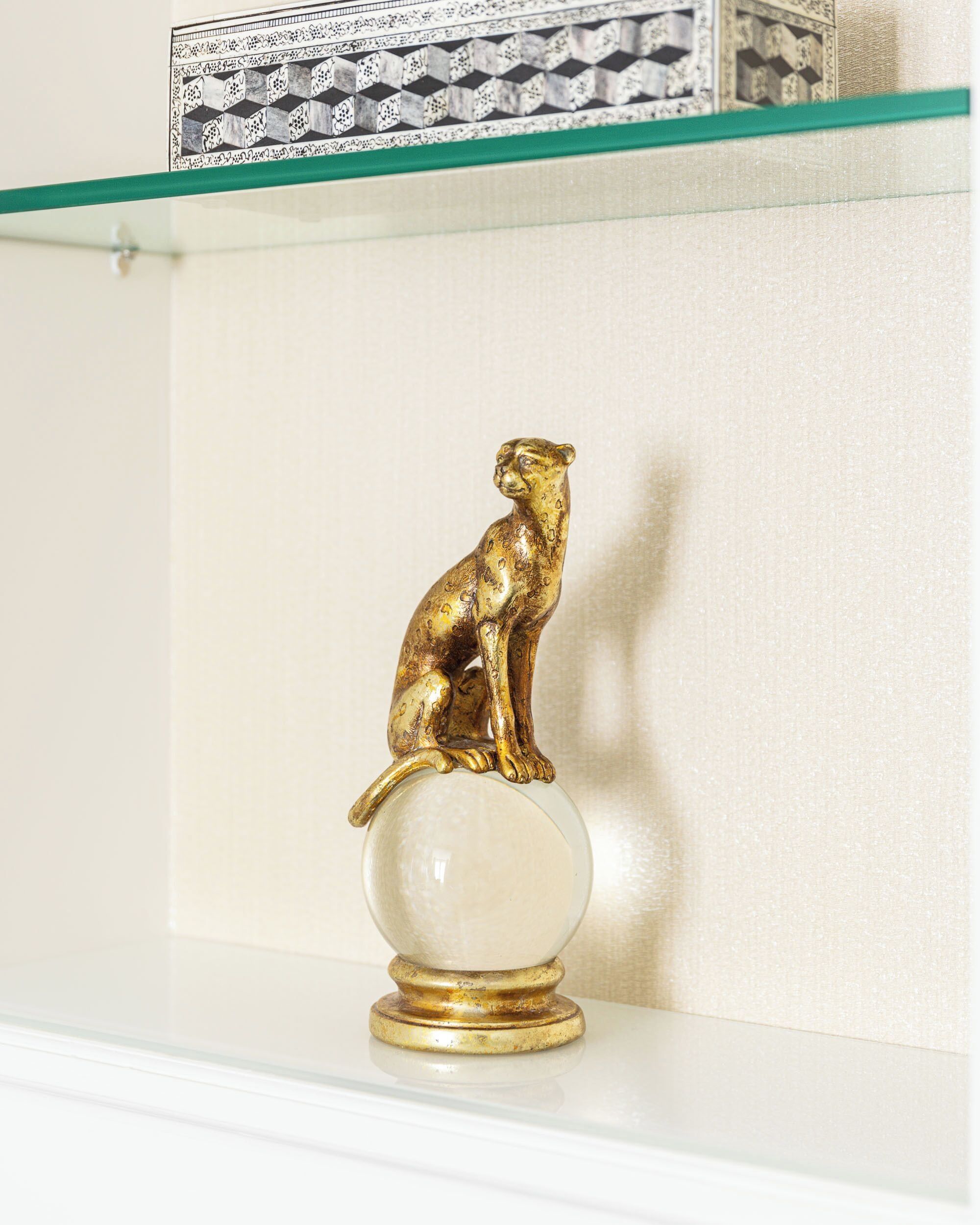 Una figurita de animal dorado sobre una esfera de vidrio transparente, colocada en un estante blanco frente a una pared lisa.