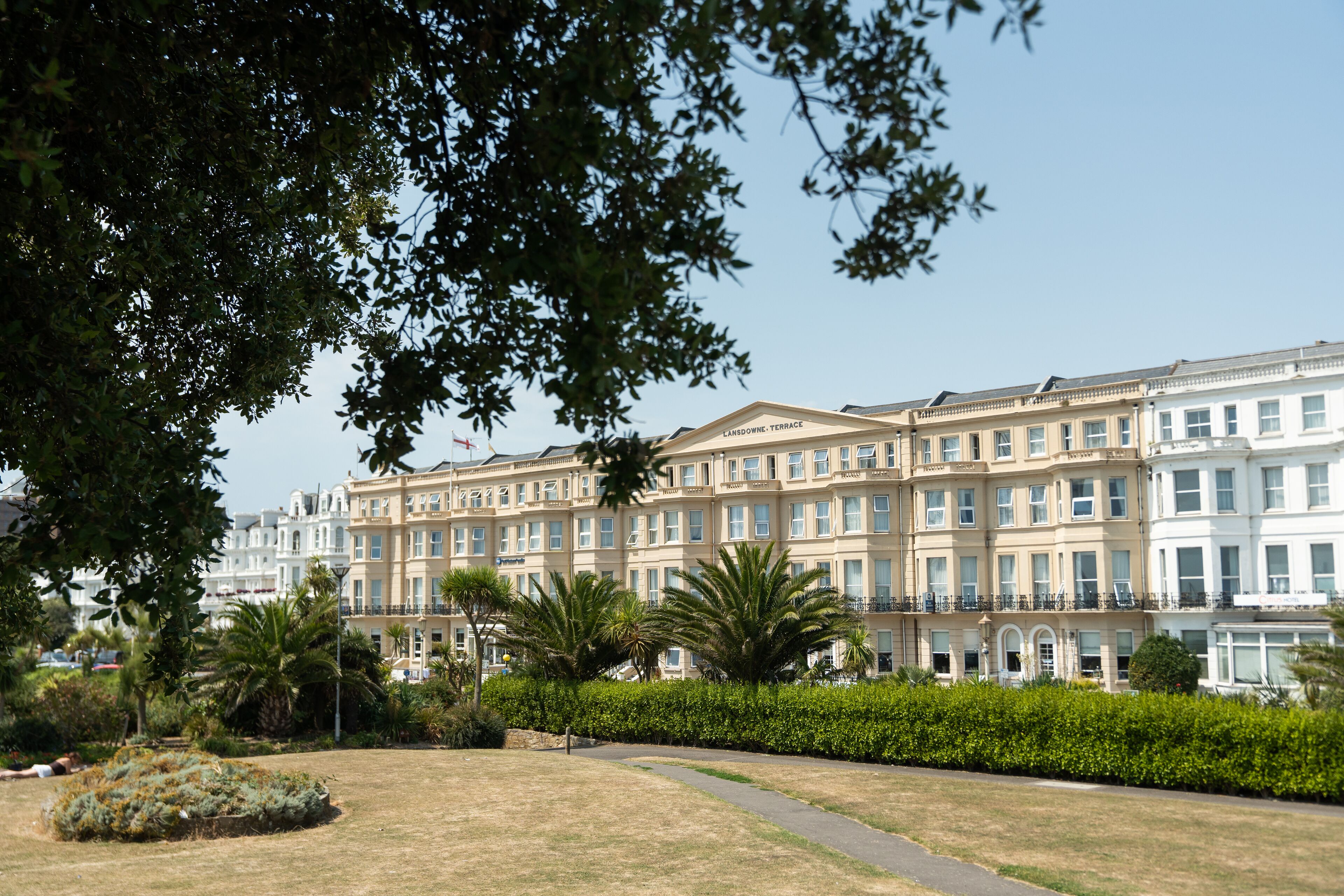 Een rij gelijkmatige, meerlaagse gebouwen in klassieke stijl, vermoedelijk woningen of hotelkamers, omgeven door verzorgde gazons en palmbomen.