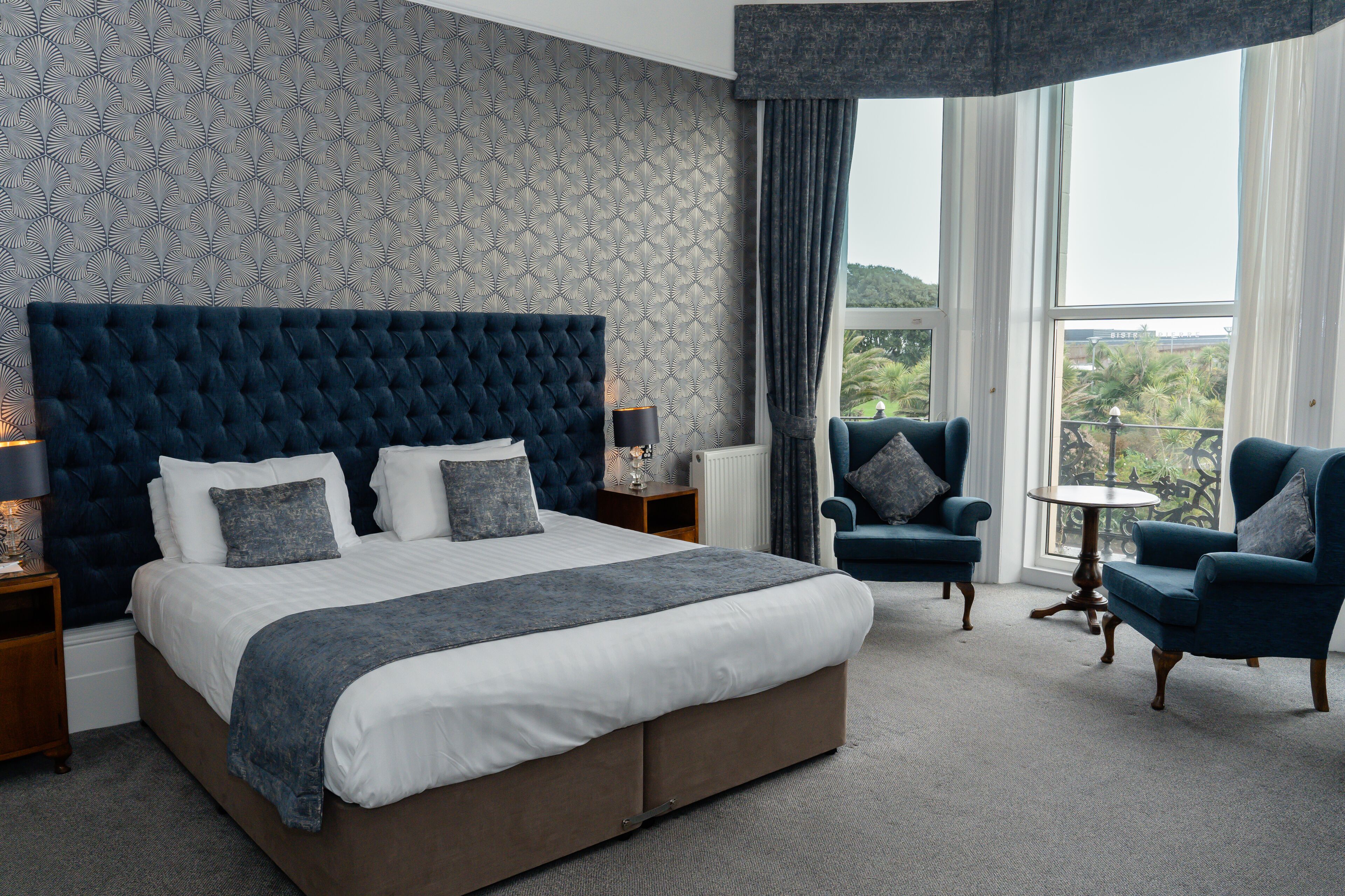 A bedroom with a large bed, two armchairs, a small table, and a view of greenery through the window.
