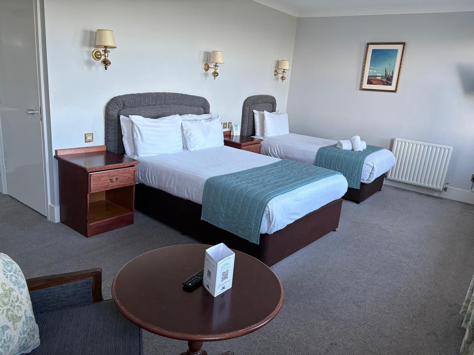 Habitación de hotel con dos camas, una mesita pequeña y una silla. Hay lámparas de pared y un cuadro enmarcado.