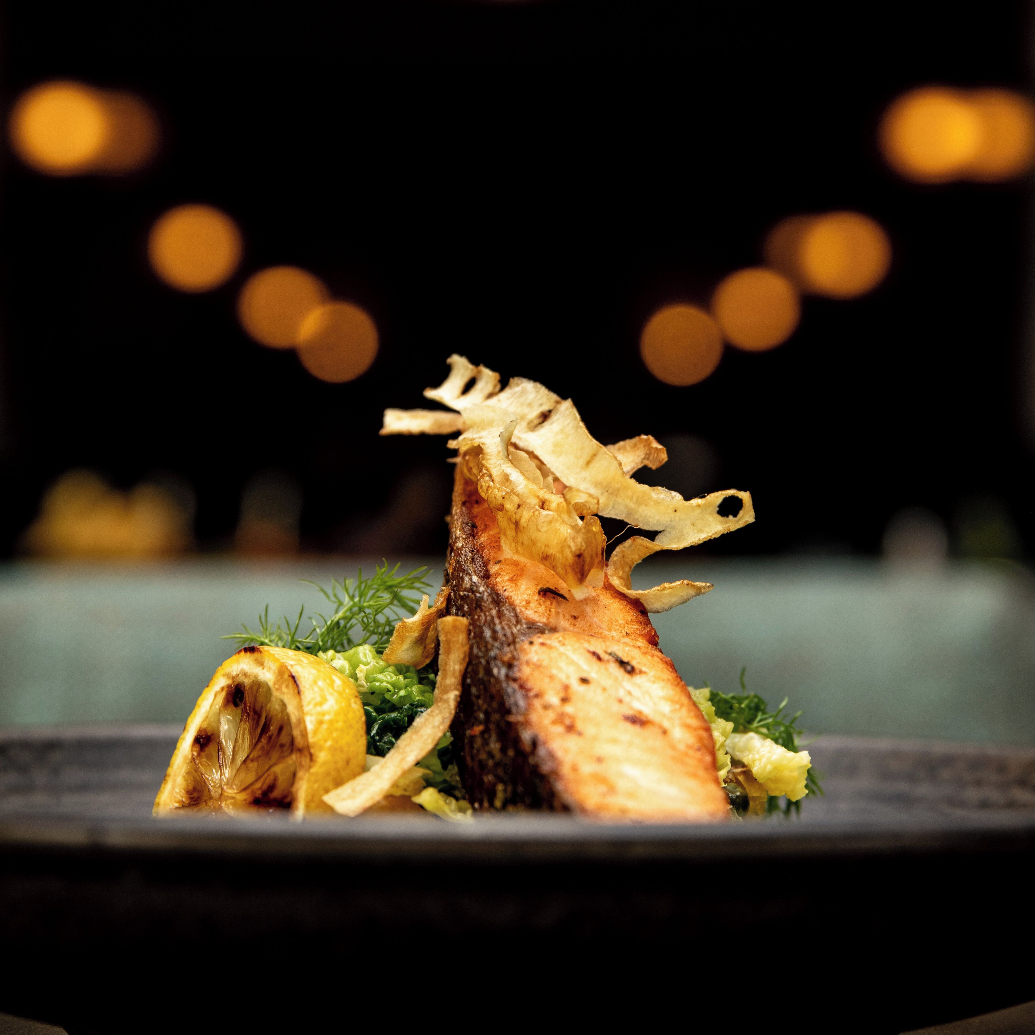 A plated dish with a grilled fish centerpiece, garnished with herbs and lemon slices.