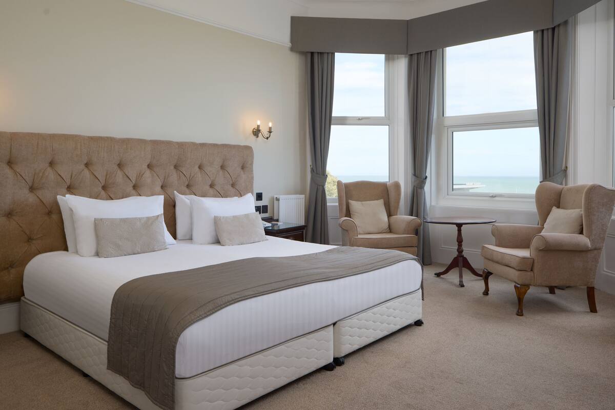 Una cama grande con cabecera capitonada, dos sillones, una mesita y vista al mar a través de amplias ventanas.
