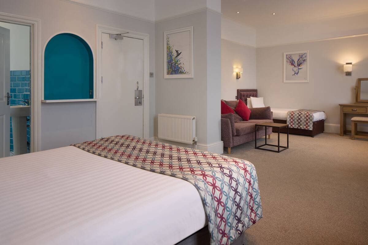 Habitación de hotel con una cama grande, un sofá, una mesita pequeña y una silla. Hay una puerta, un radiador y obras de arte en las paredes.